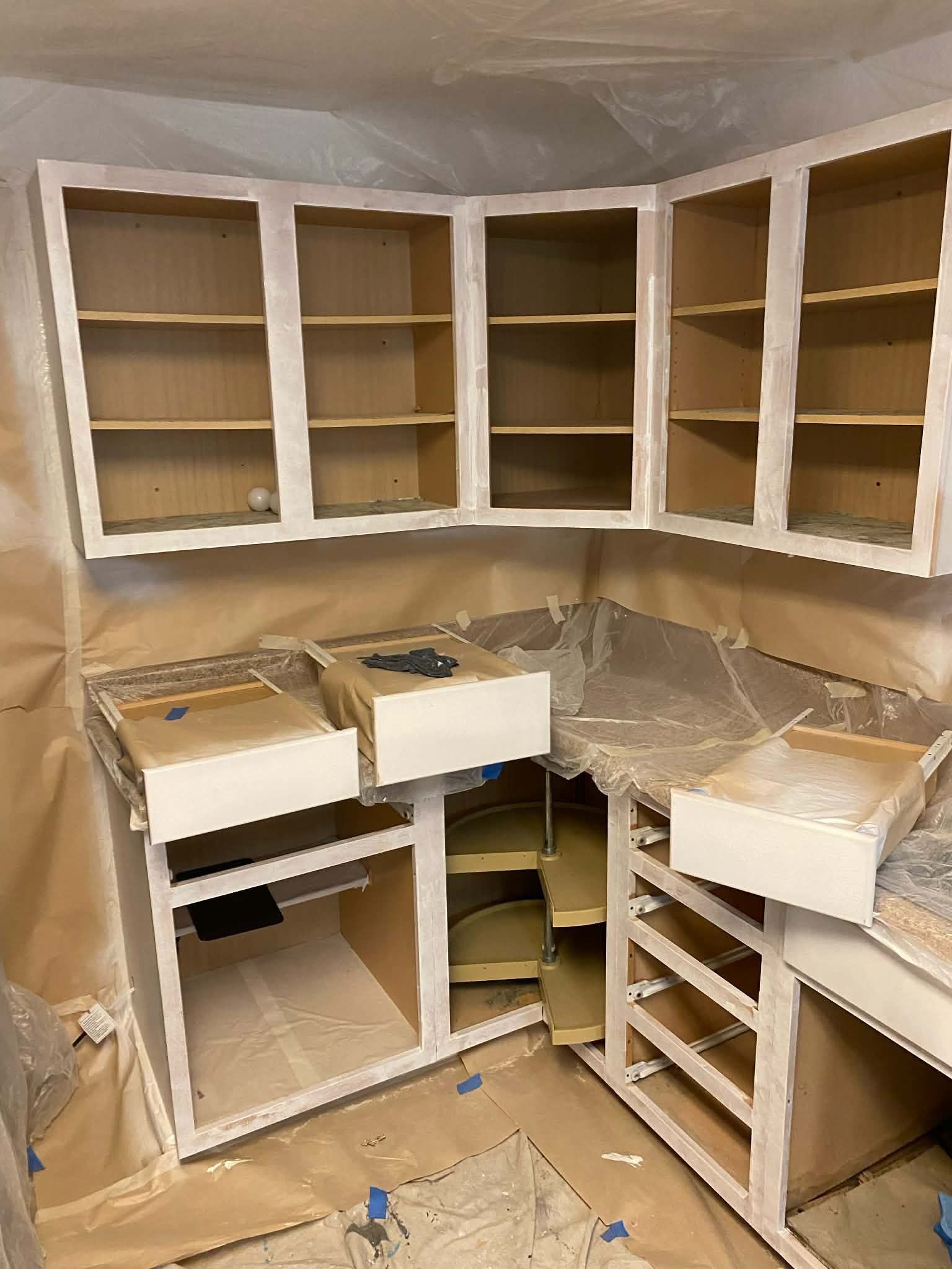 Kitchen cabinets in the process of being painted white, with walls and surfaces covered in protective paper.