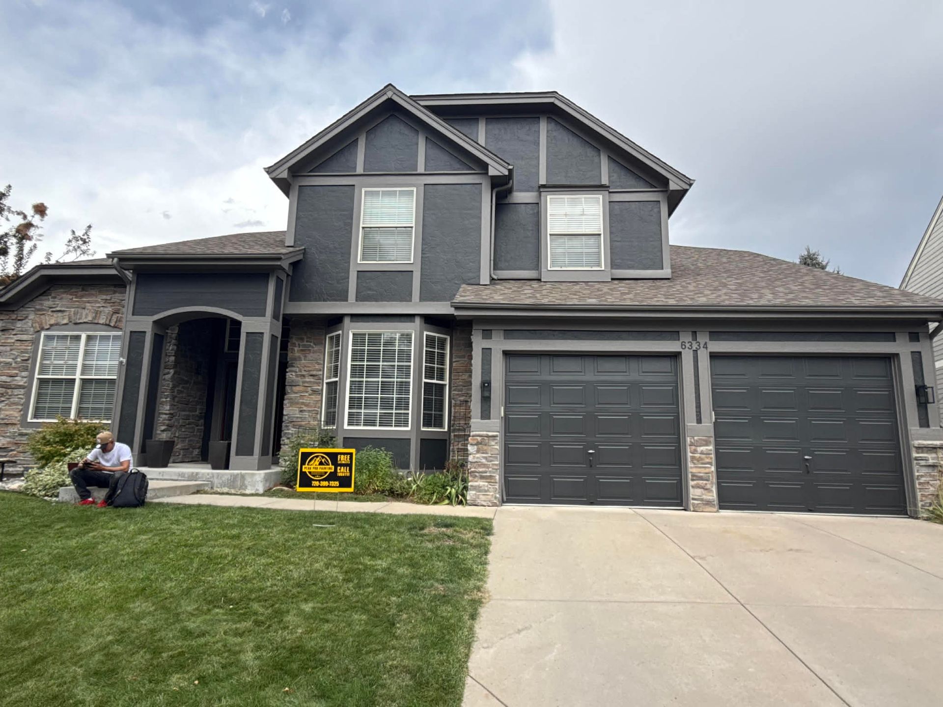 A two-story house with dark gray siding, stone accents, and a two-car garage, with a person sitting on the lawn.