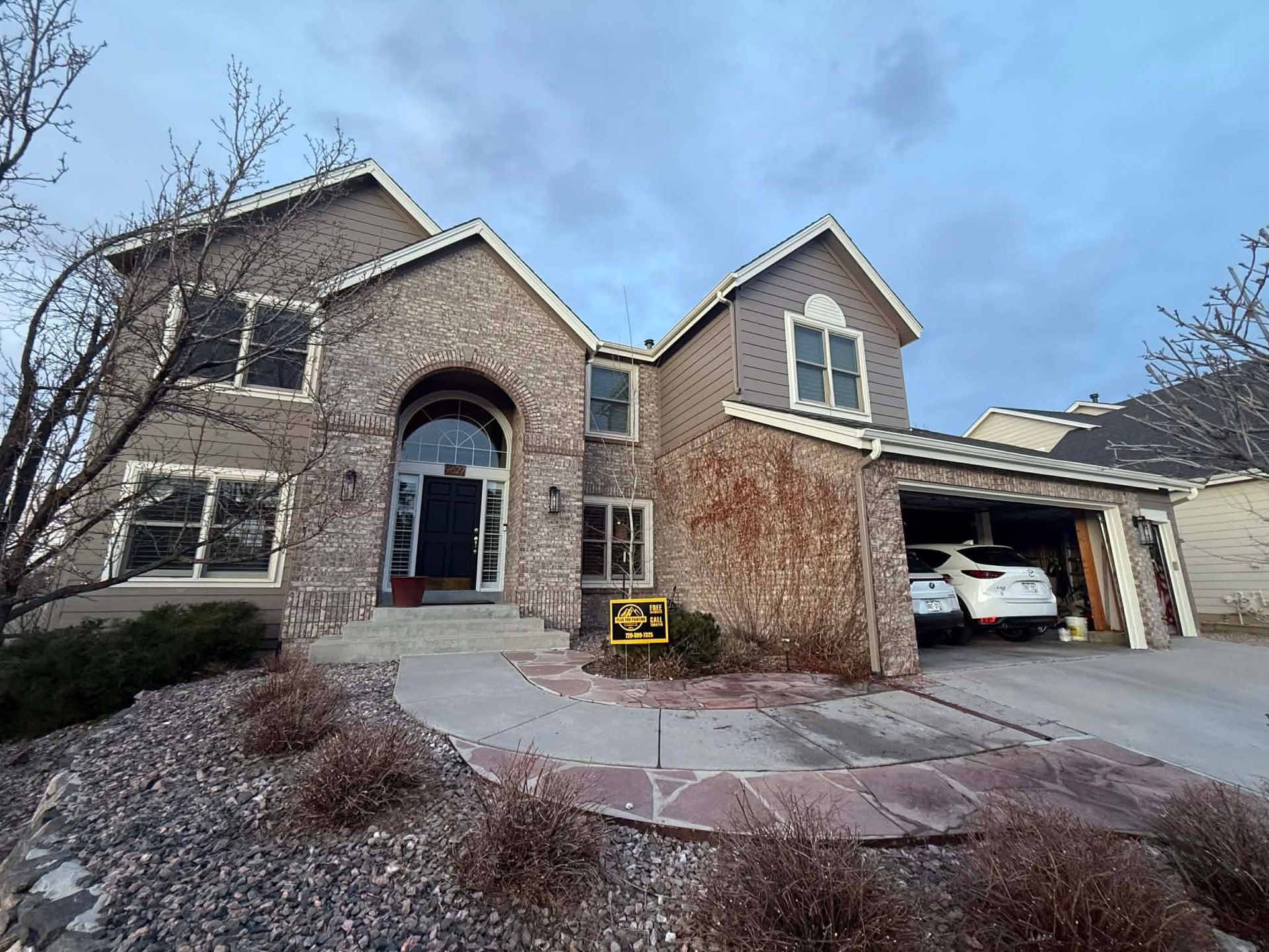 A two-story tan brick house with a arched entryway, a attached two-car garage, and a flagstone walkway.