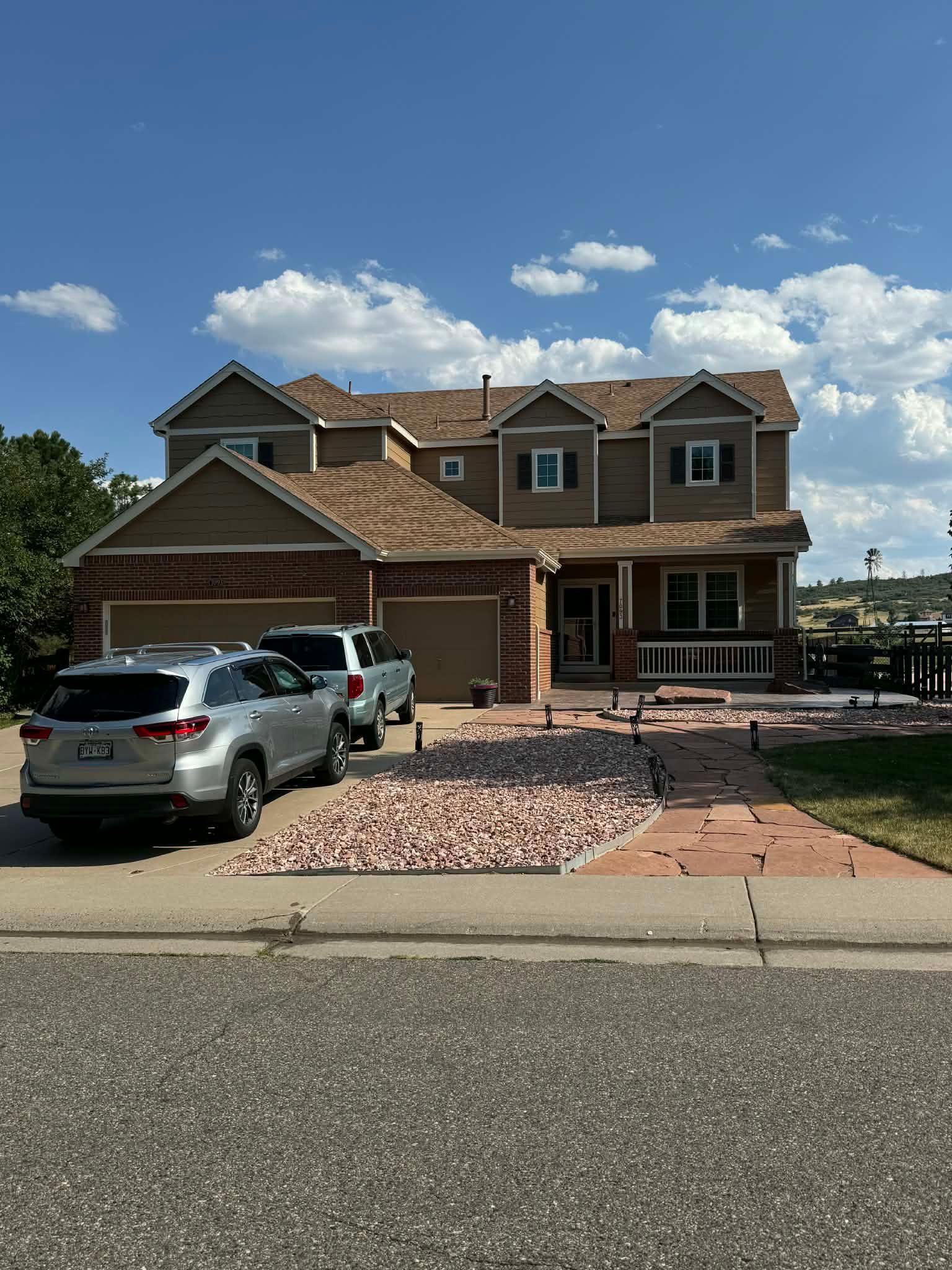 A two-story tan suburban home with a brick-front garage, two cars in the driveway, and a stone-paved walkway.