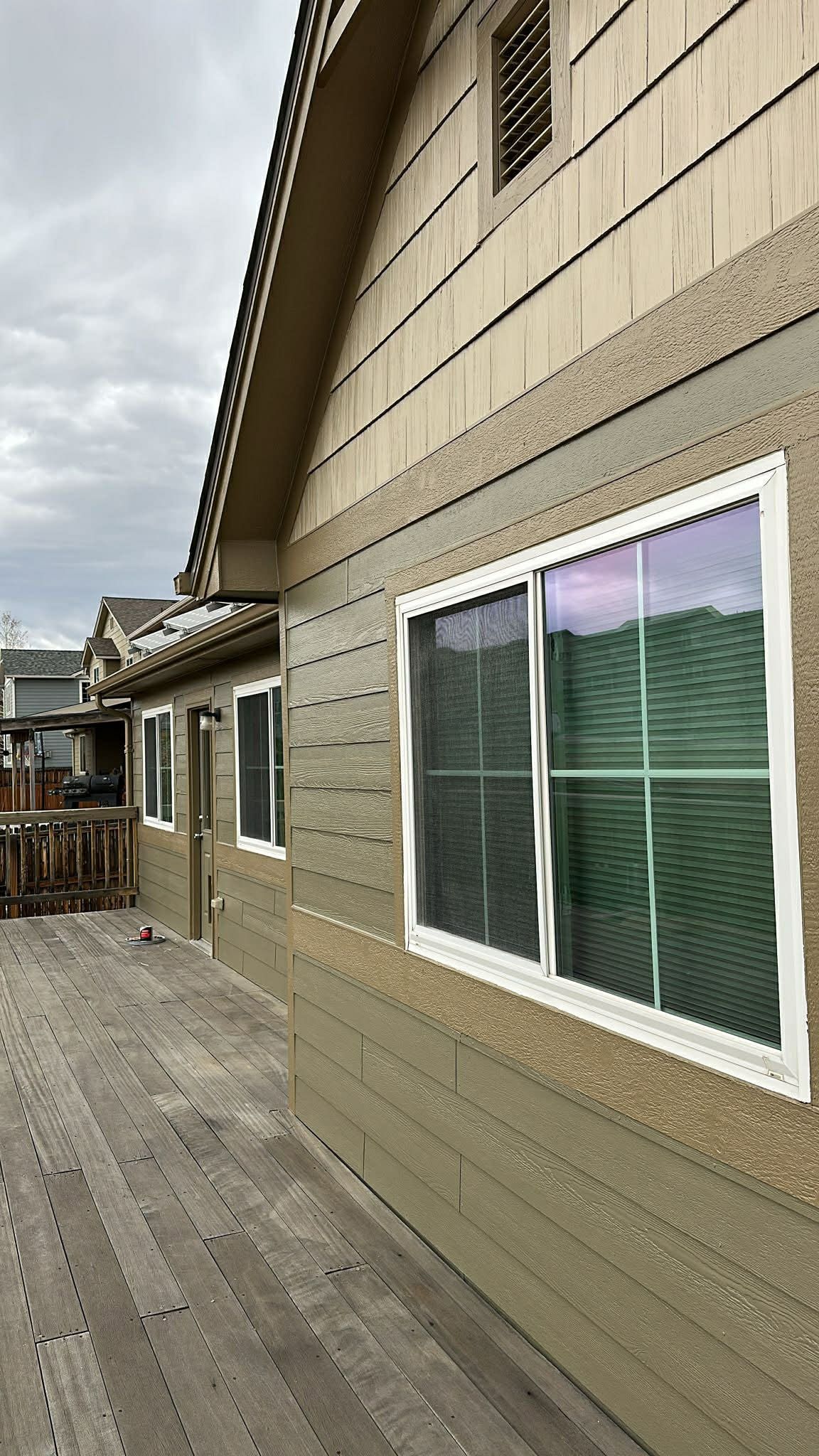 A wooden deck leads along the side of a tan house with a large window and textured siding under a cloudy sky.