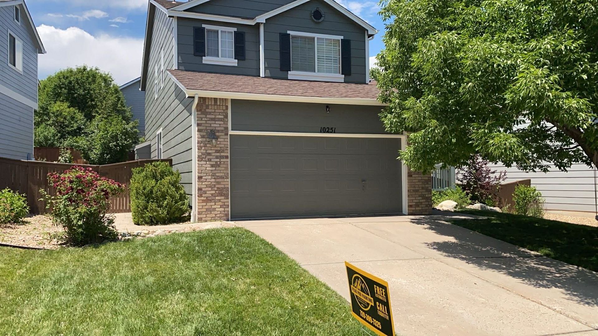 A two-story grey suburban house with a brick accent, a large garage, a green lawn, and a sign in the driveway.