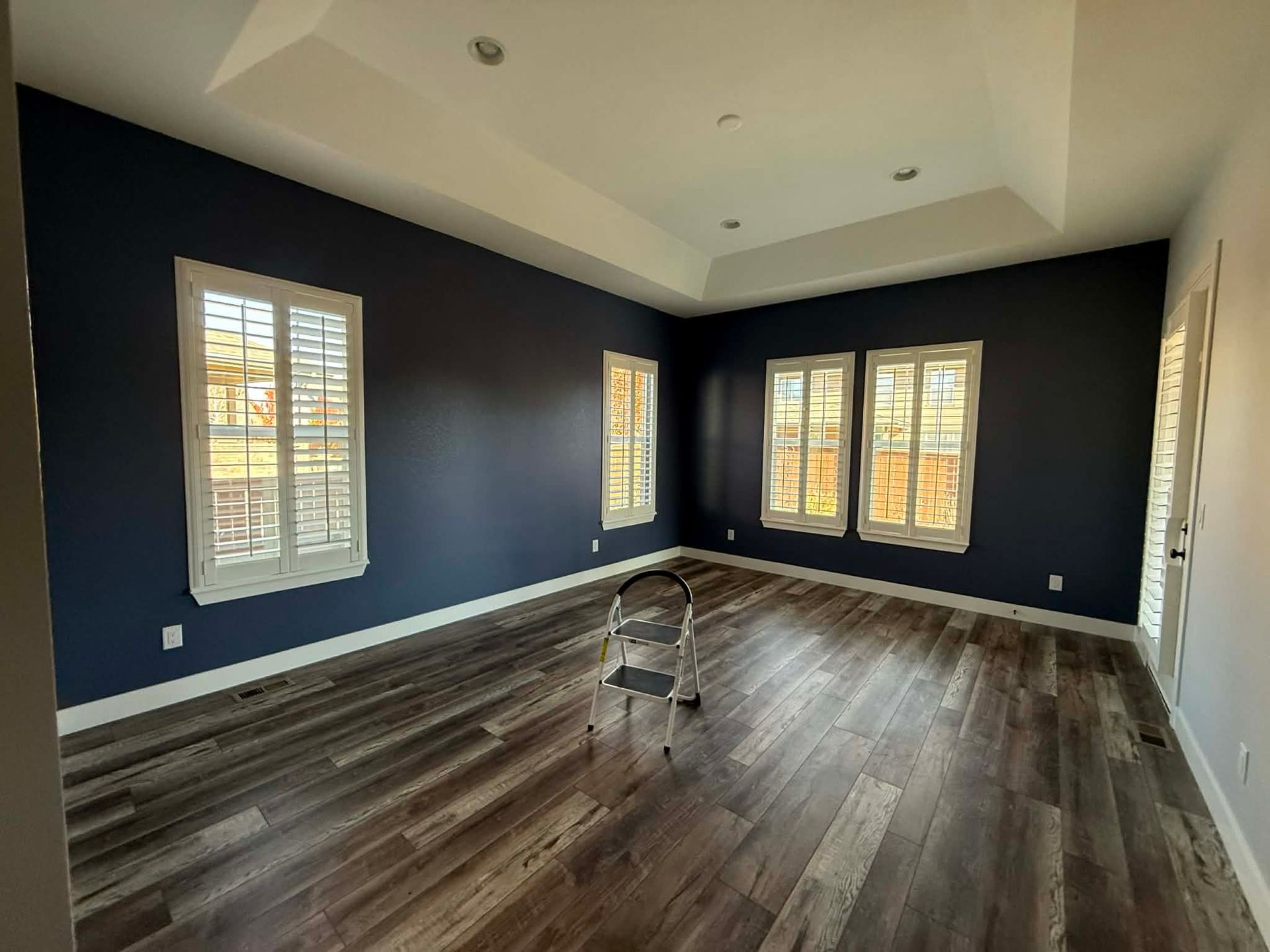 An empty room with dark blue walls, wood-look flooring, white plantation shutters, and a central folding step stool.