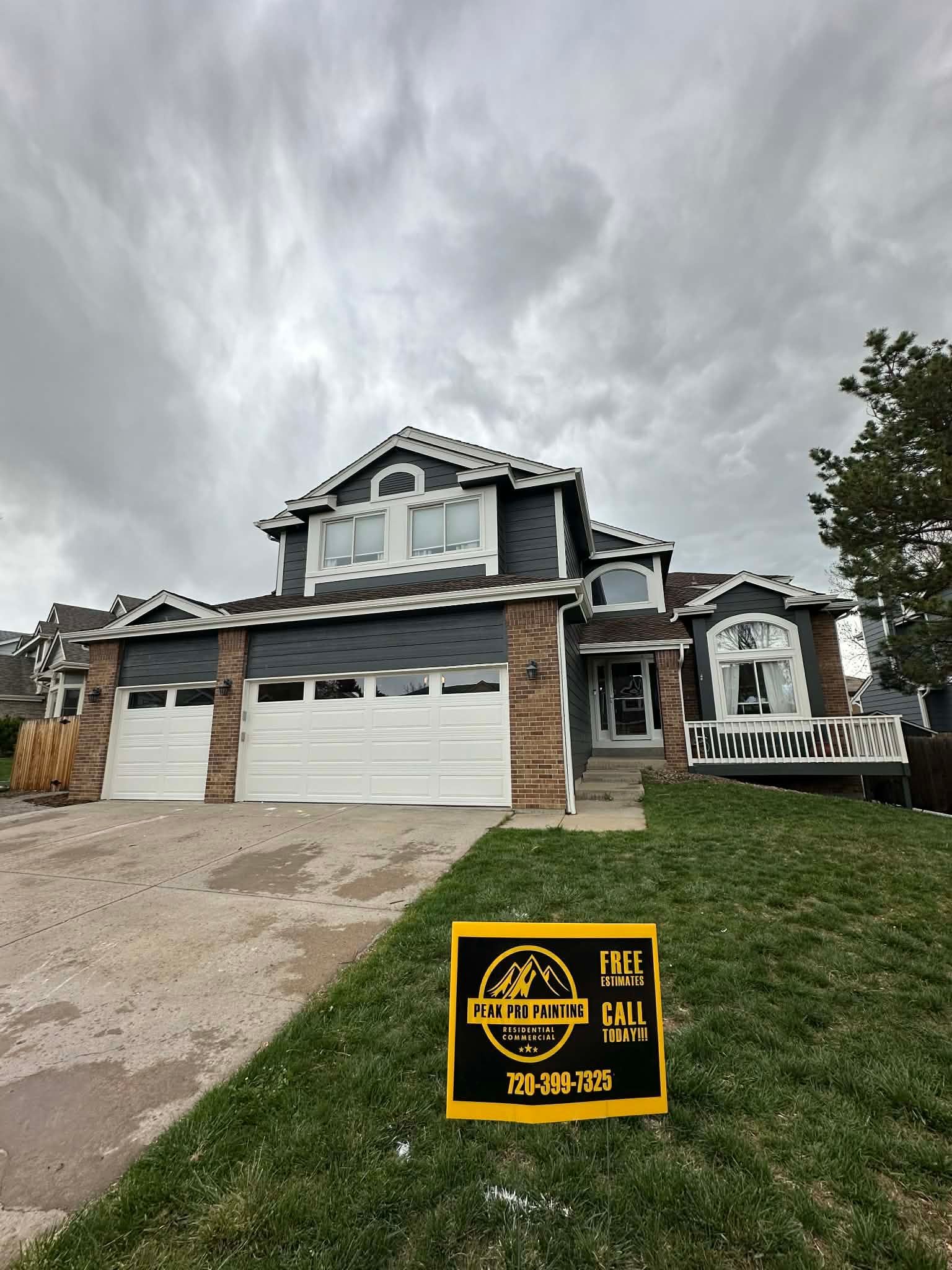 A two-story gray house with stone accents under a cloudy sky, with a yellow construction yard sign on the front lawn.