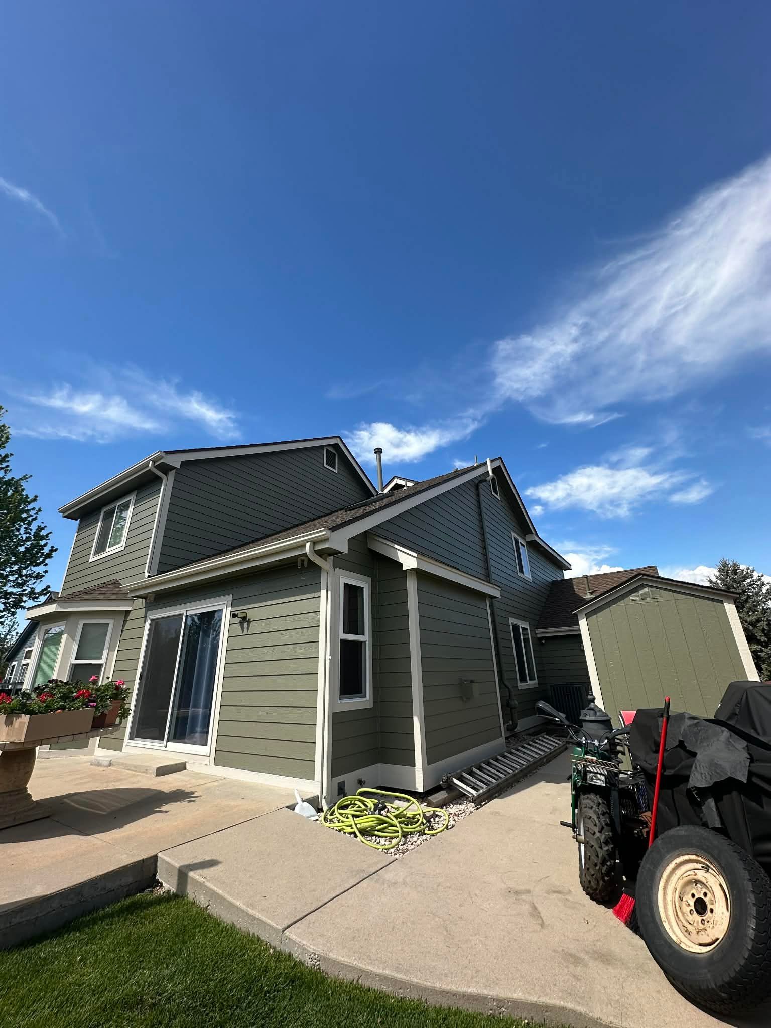 An olive-green house with white trim and a black shingled roof under a bright blue sky, with a utility trailer nearby.