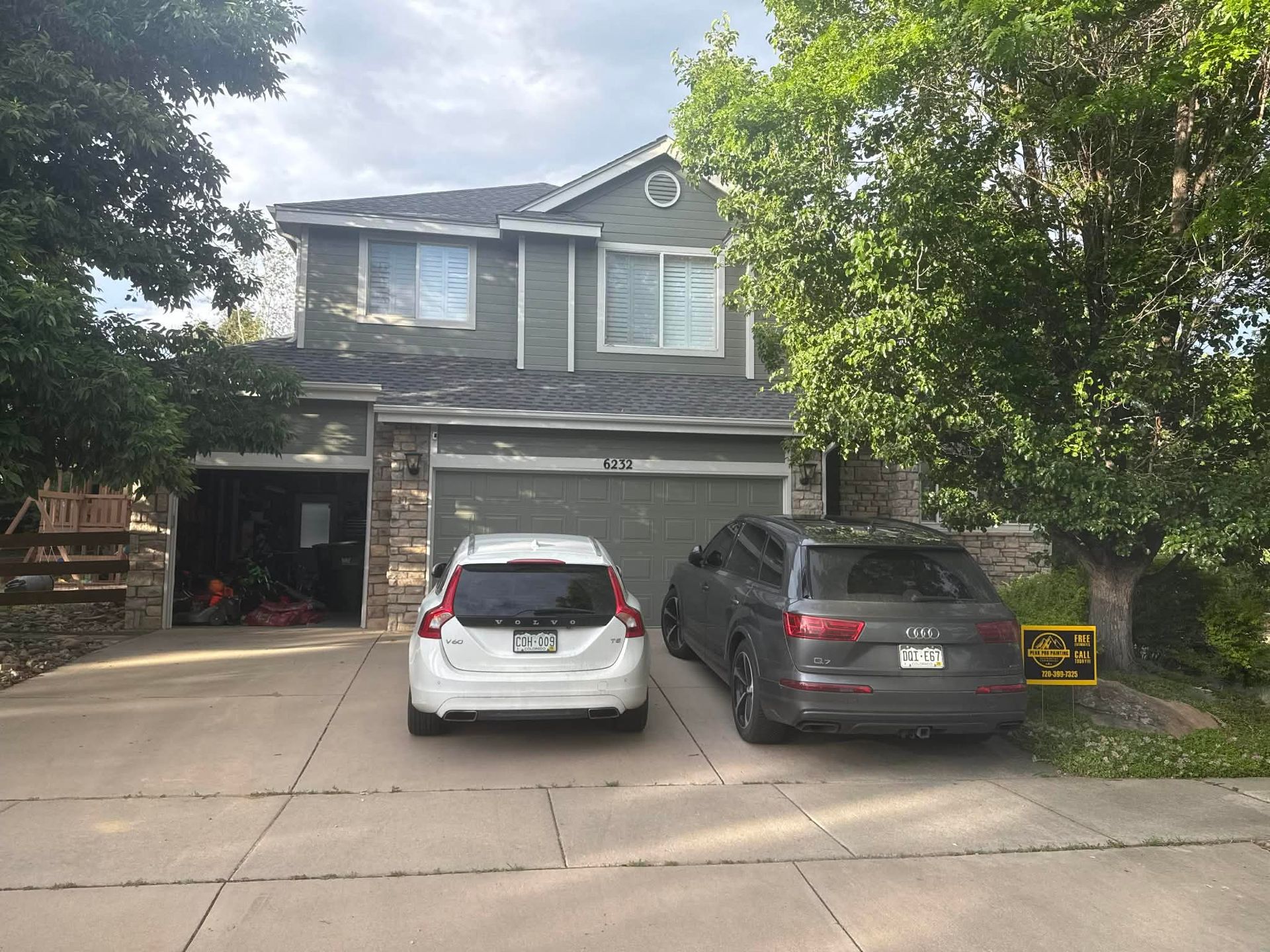 A two-story grey suburban house with a driveway holding a white SUV and a dark grey SUV under a cloudy sky.