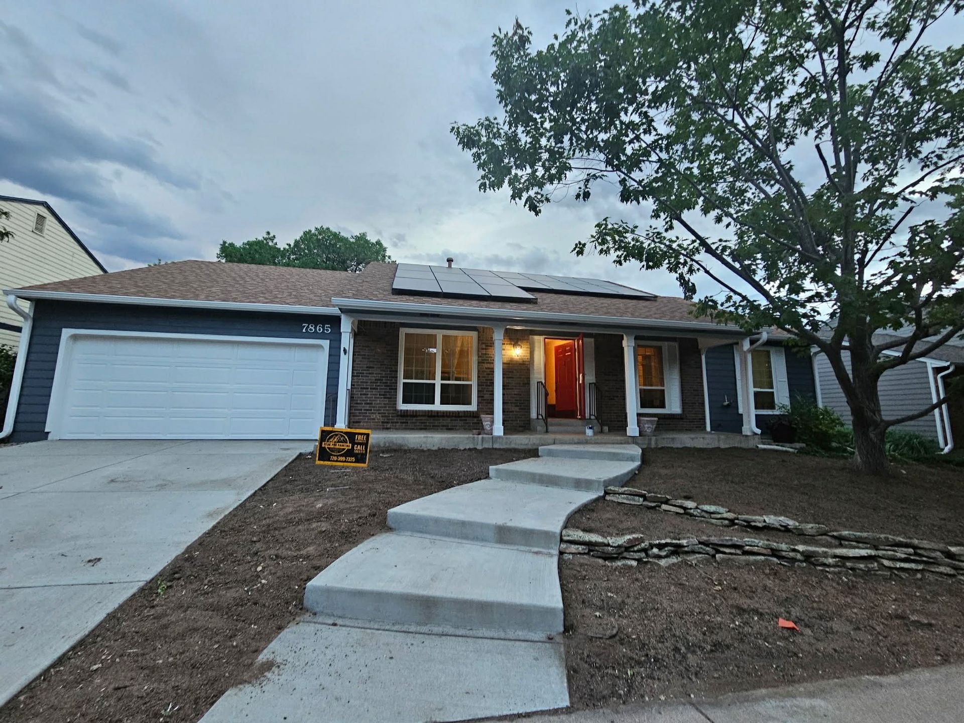 A single-story blue house with a white garage, dark stone accents, solar panels on the roof, and a concrete path entry.