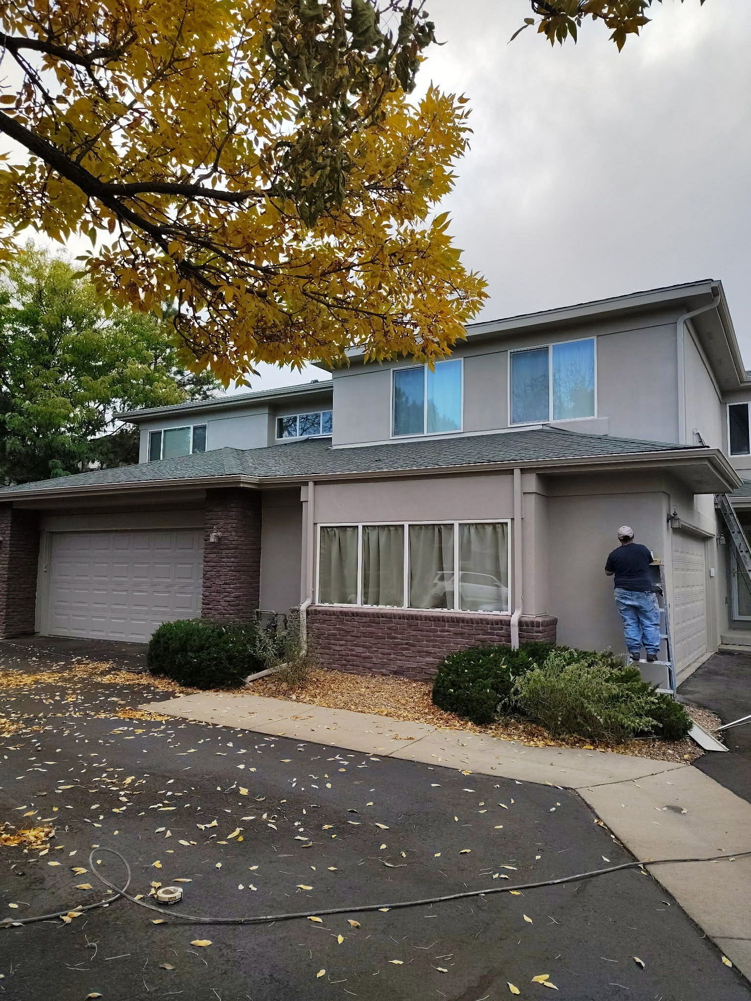 A person stands on a ladder, painting the exterior wall of a two-story gray house with a stone-accented front.