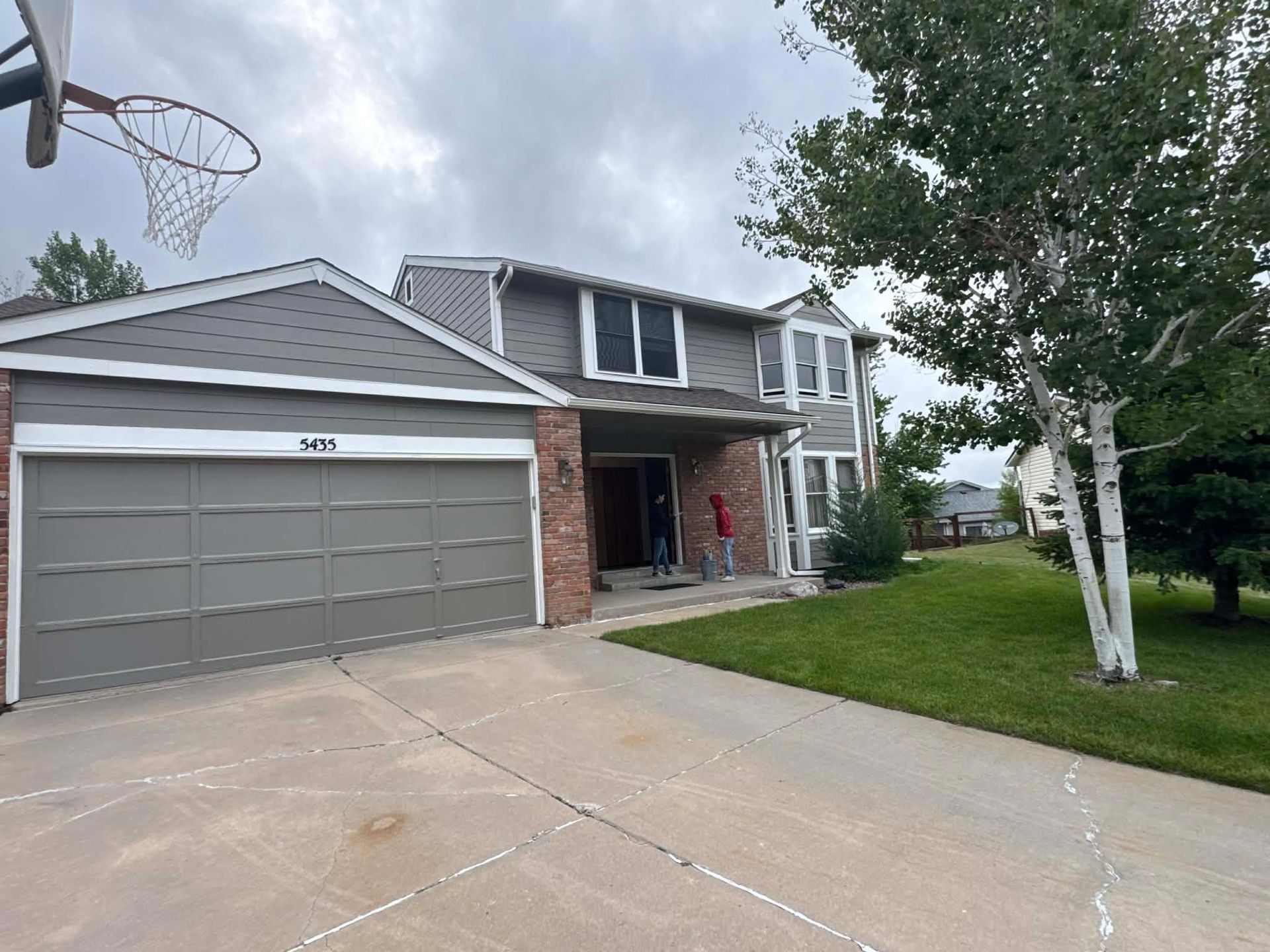 A two-story grey suburban house with a brick accent, a large garage, and a driveway with a basketball hoop on the left.