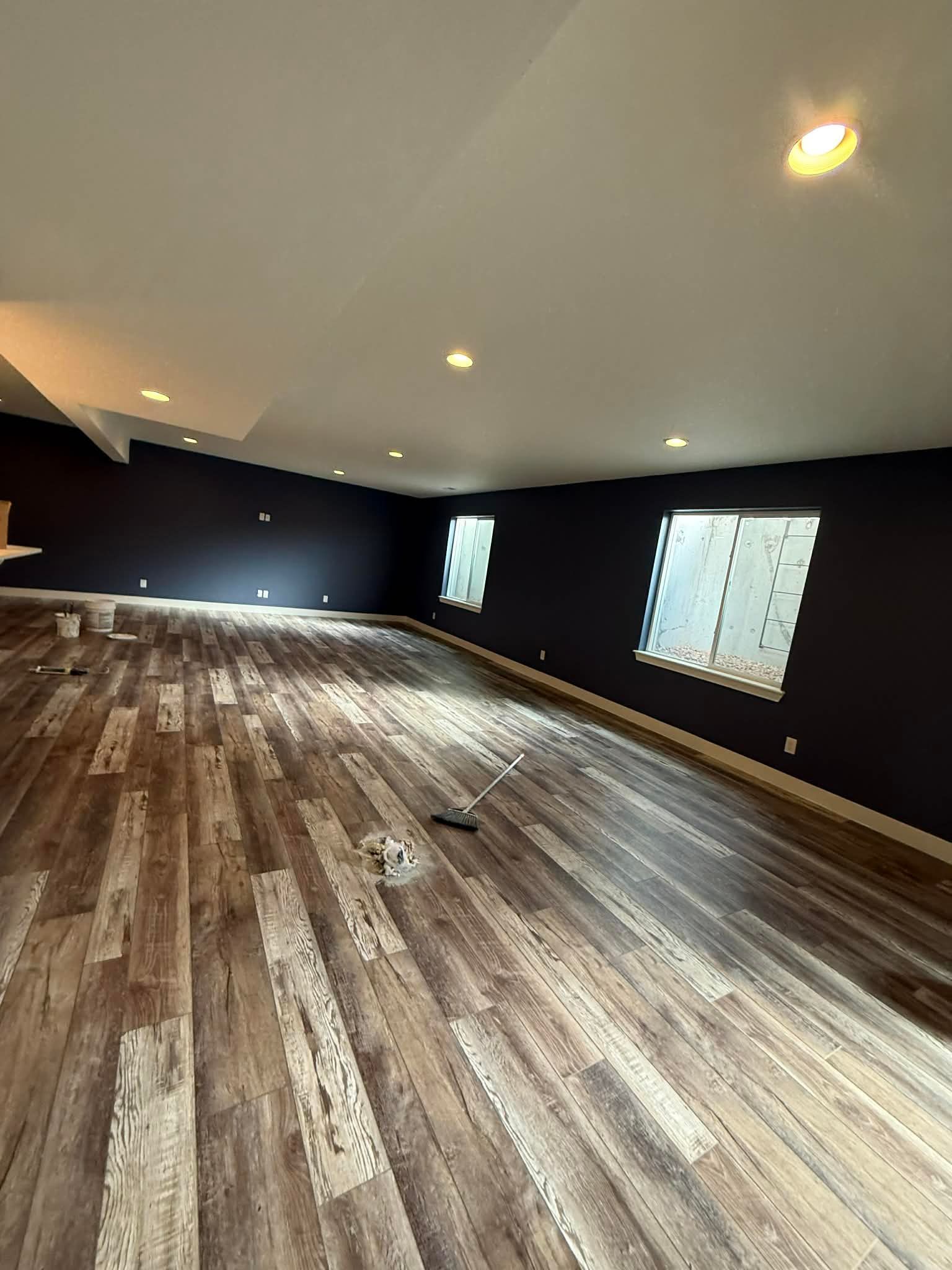 A spacious, empty room featuring dark walls, recessed ceiling lights, and light wood-plank flooring with two windows.