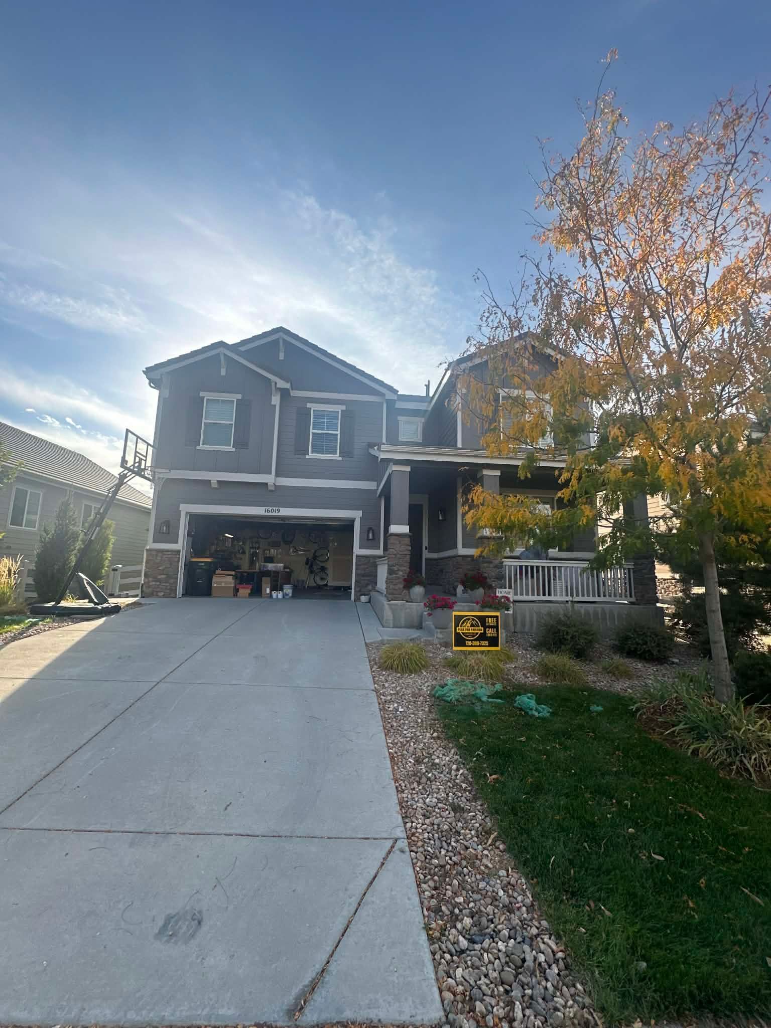 A gray two-story house with a driveway, open garage, and a large tree with autumn leaves in the front yard.
