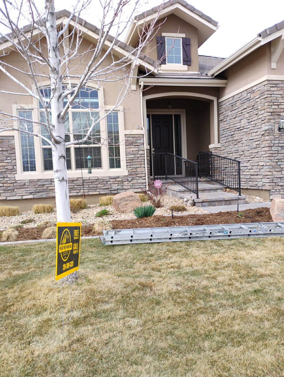 A suburban house with stone siding and tan trim, featuring a front porch, a birch tree, and a yard sign in the grass.