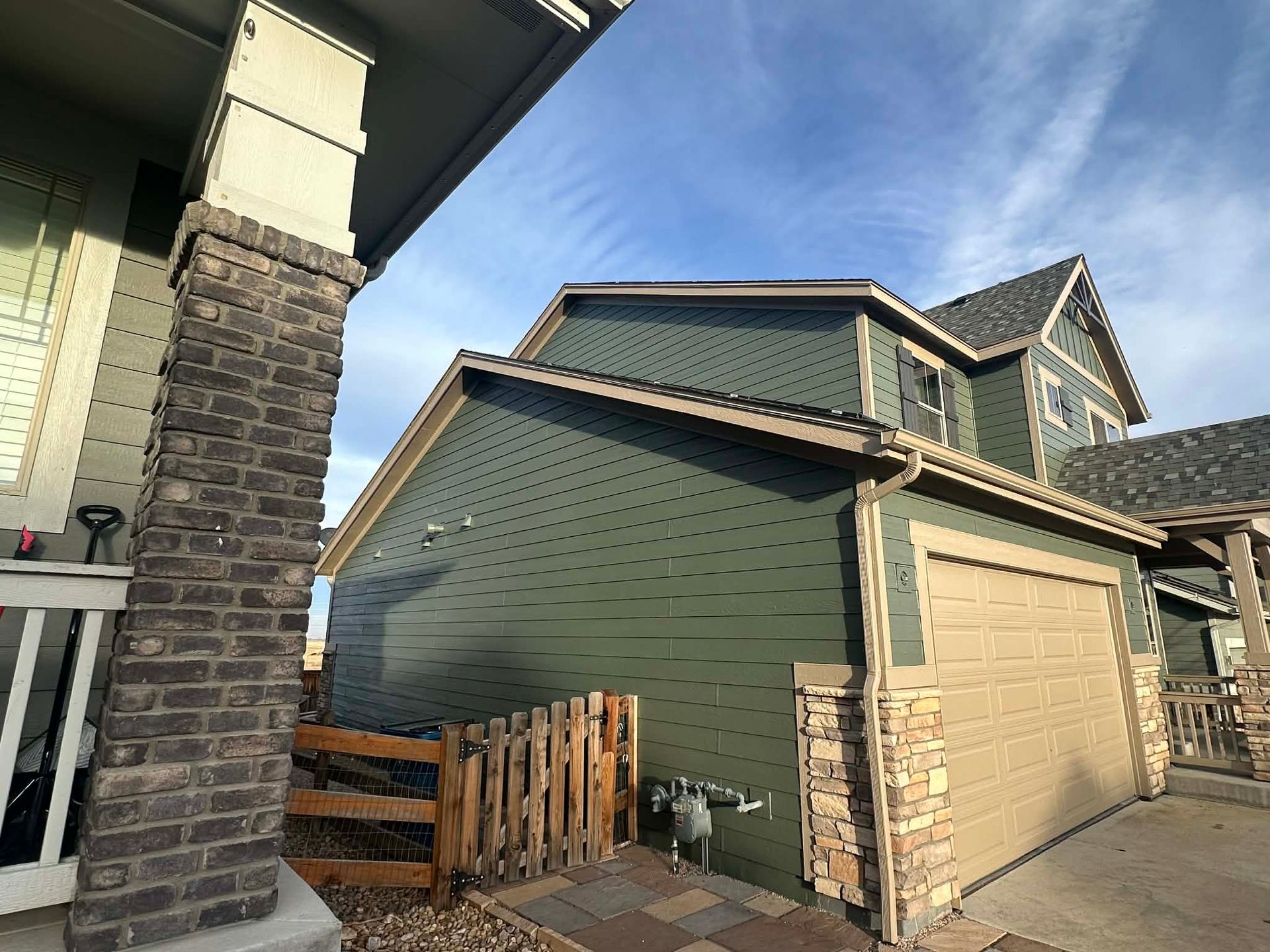 A green house with a tan garage door and stone pillars under a blue sky, viewed from a porch perspective.