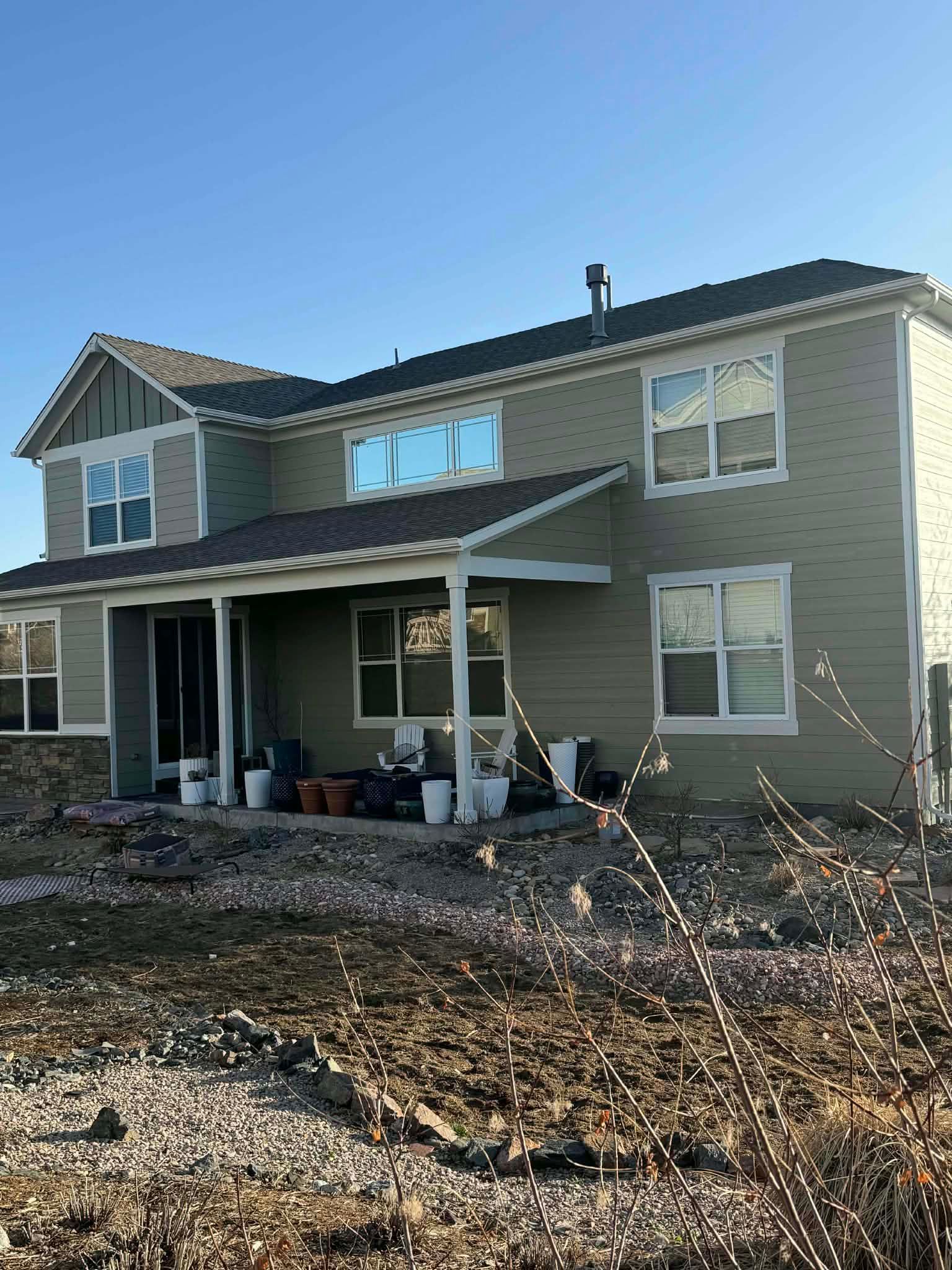 A two-story green house with a covered front porch, white trim, and a gravel yard under a clear blue sky.