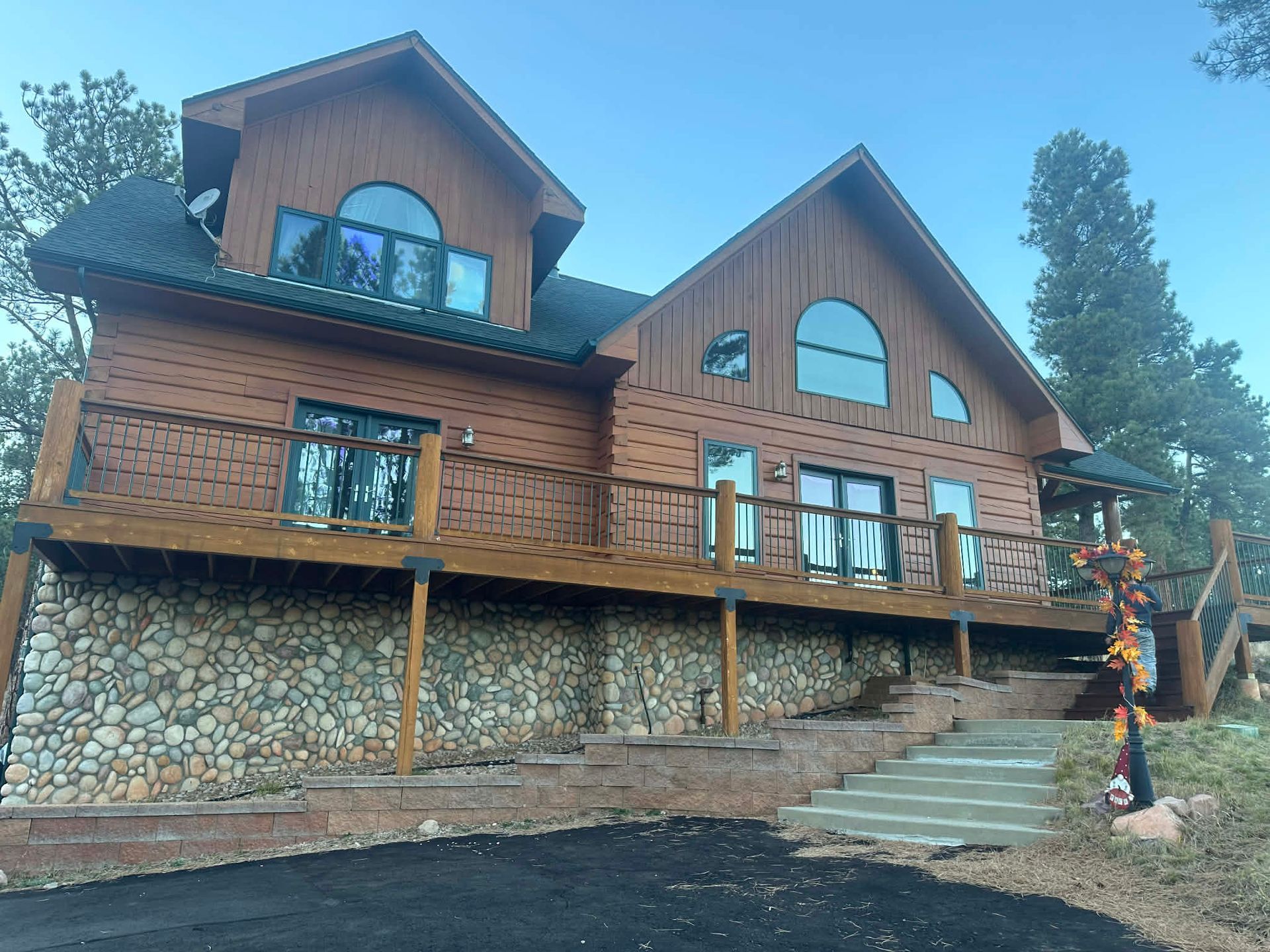 A two-story wood-sided cabin with a stone foundation, large wooden deck, and green roof in a forest setting.