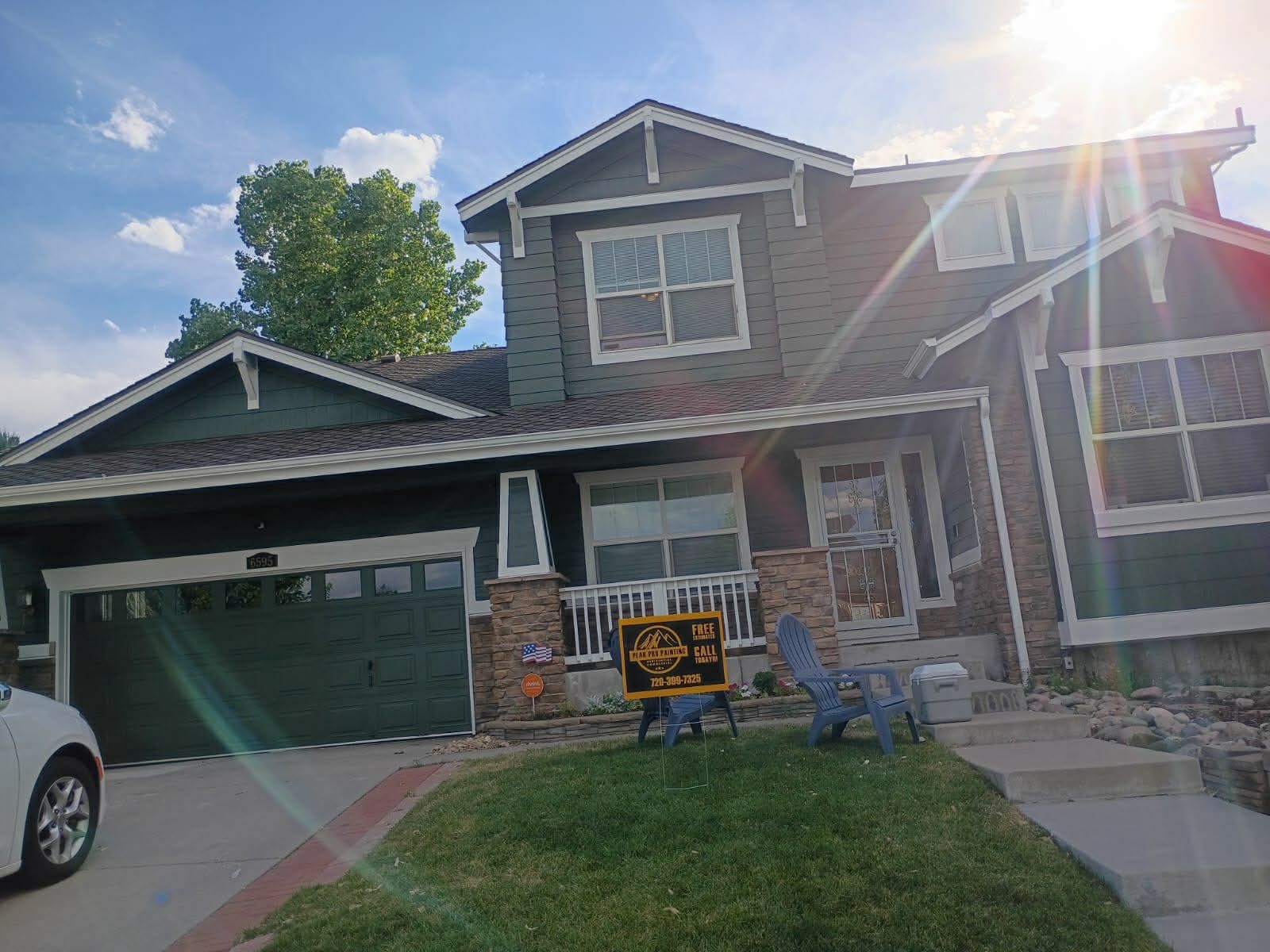 A two-story dark green home with a garage, stone accents, and a front yard featuring blue lawn chairs and a small sign.