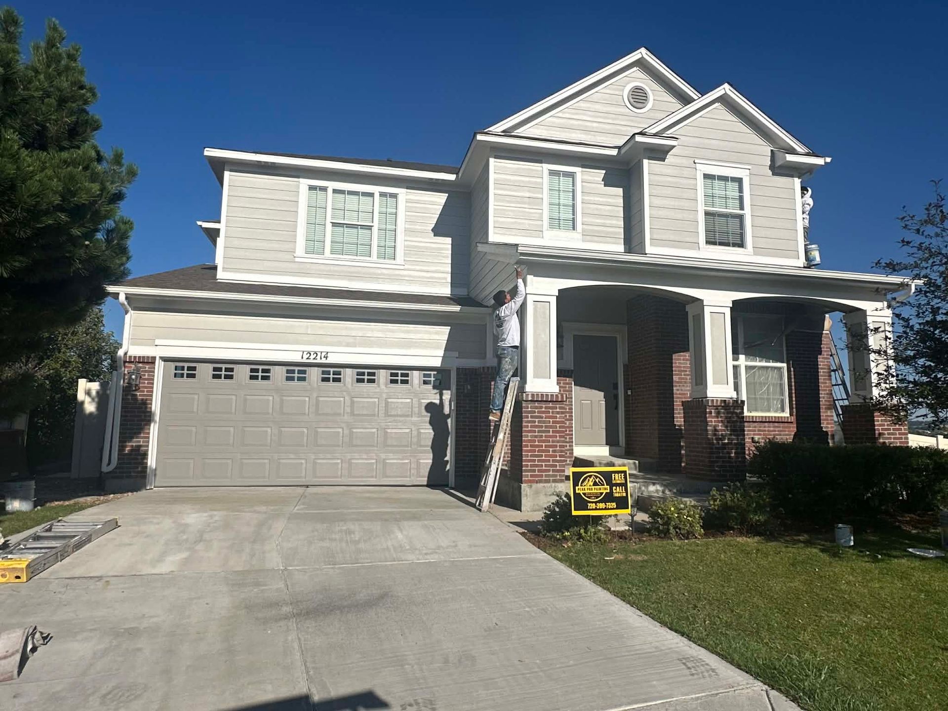 A person on a ladder paints the exterior trim of a two-story beige house with a brick base and a gray garage door.