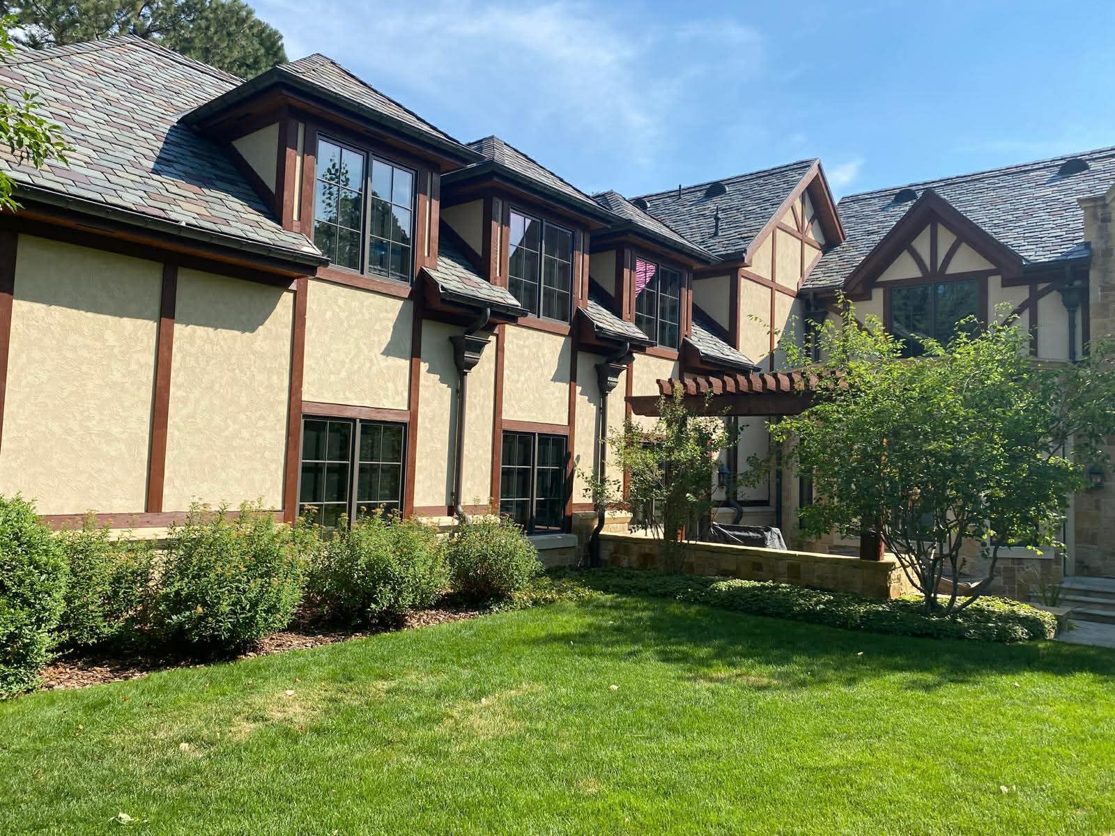 A multi-story Tudor-style home with light-colored stucco and brown timber framing, surrounded by a green lawn and trees.