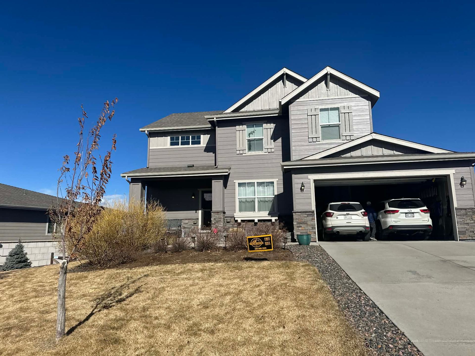 A two-story grey suburban house with a two-car garage and a dry lawn under a clear blue sky.