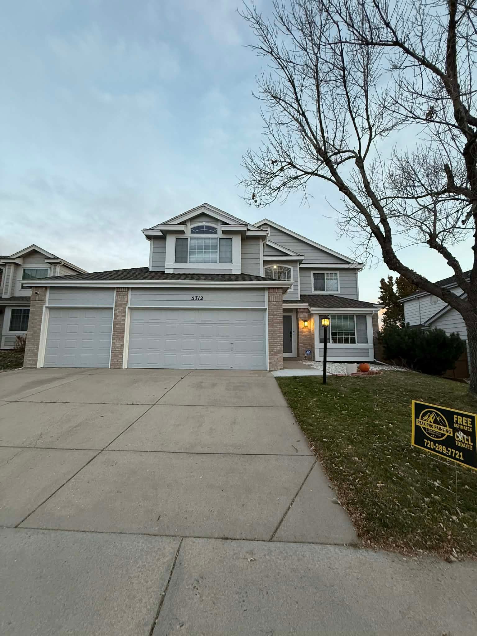 A light-colored, two-story suburban house with a three-car garage and a grassy front yard under a cloudy sky.