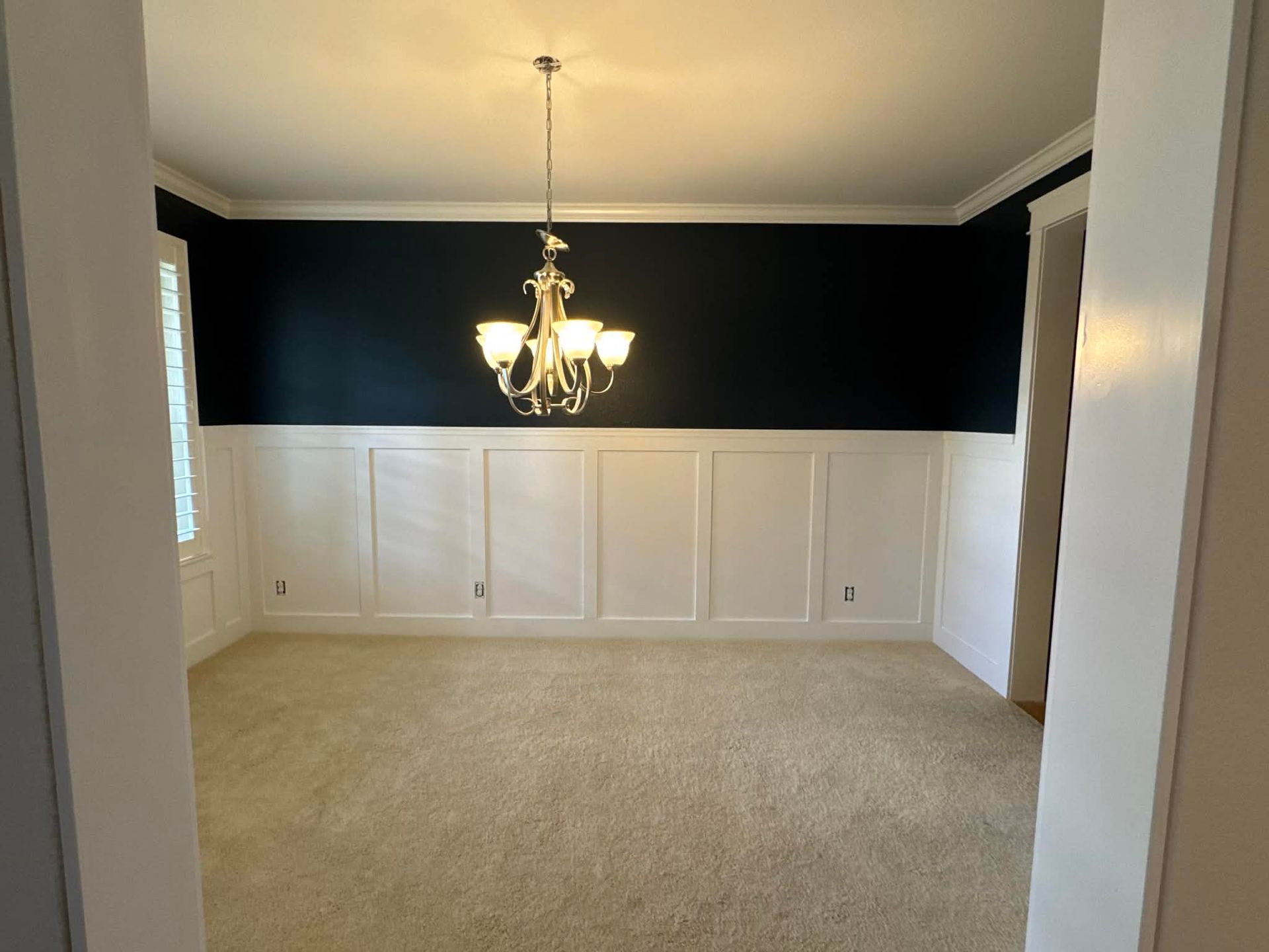 A dining room with white wainscoting on the lower walls, dark navy upper walls, beige carpeting, and a gold chandelier.
