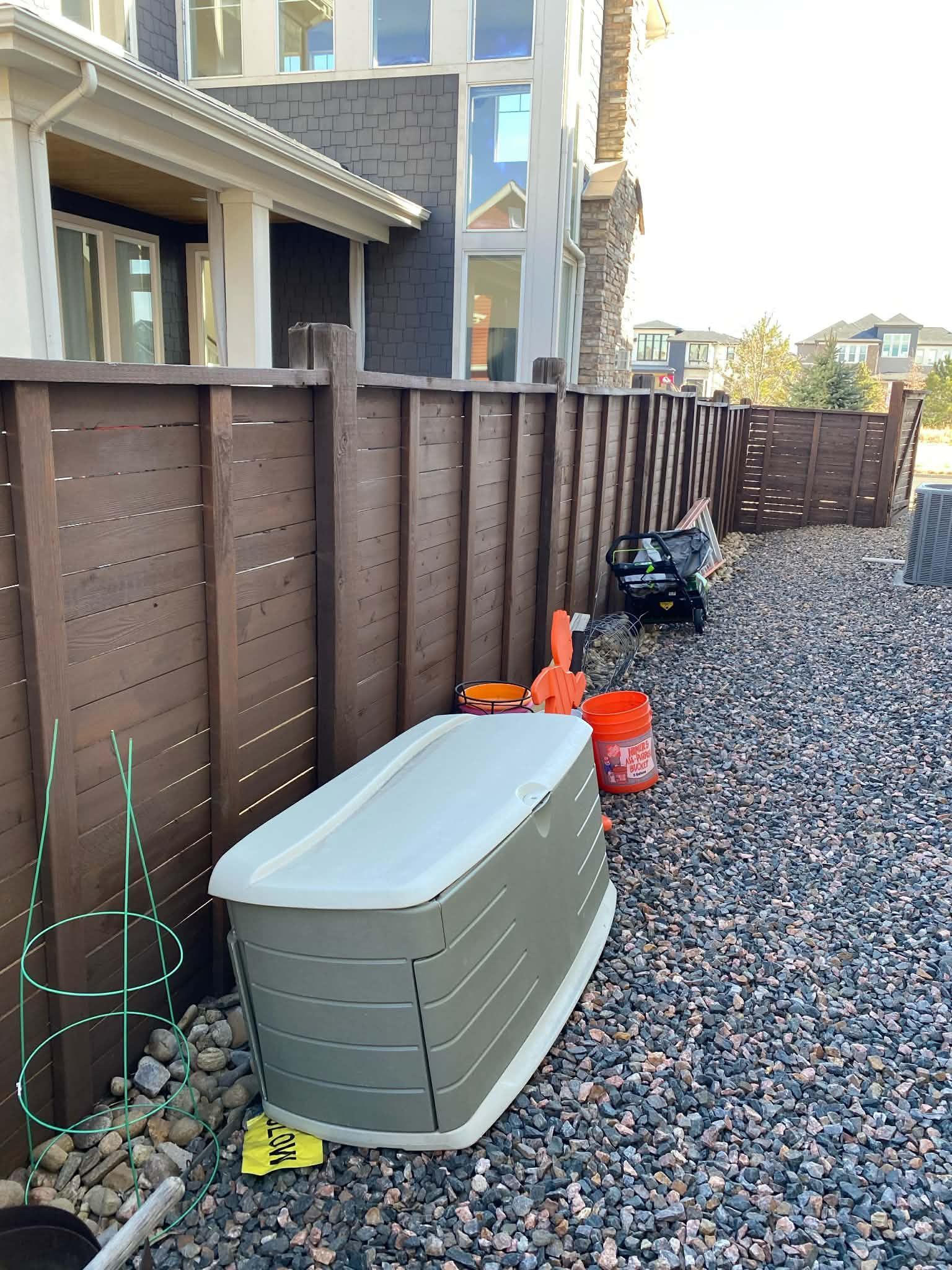 A large, horizontal, light-grey storage bin sits on a gravel yard next to a brown wooden fence behind a house.