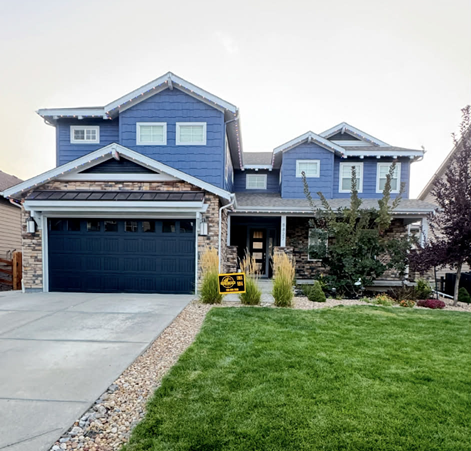 A two-story blue house with stone siding, a dark garage, and a green front lawn under a light sky.