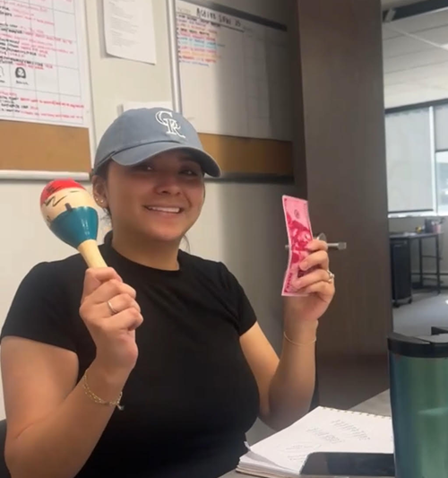 A smiling person wears a blue baseball cap, holding a maraca in one hand and a pink bill in the other at a desk.