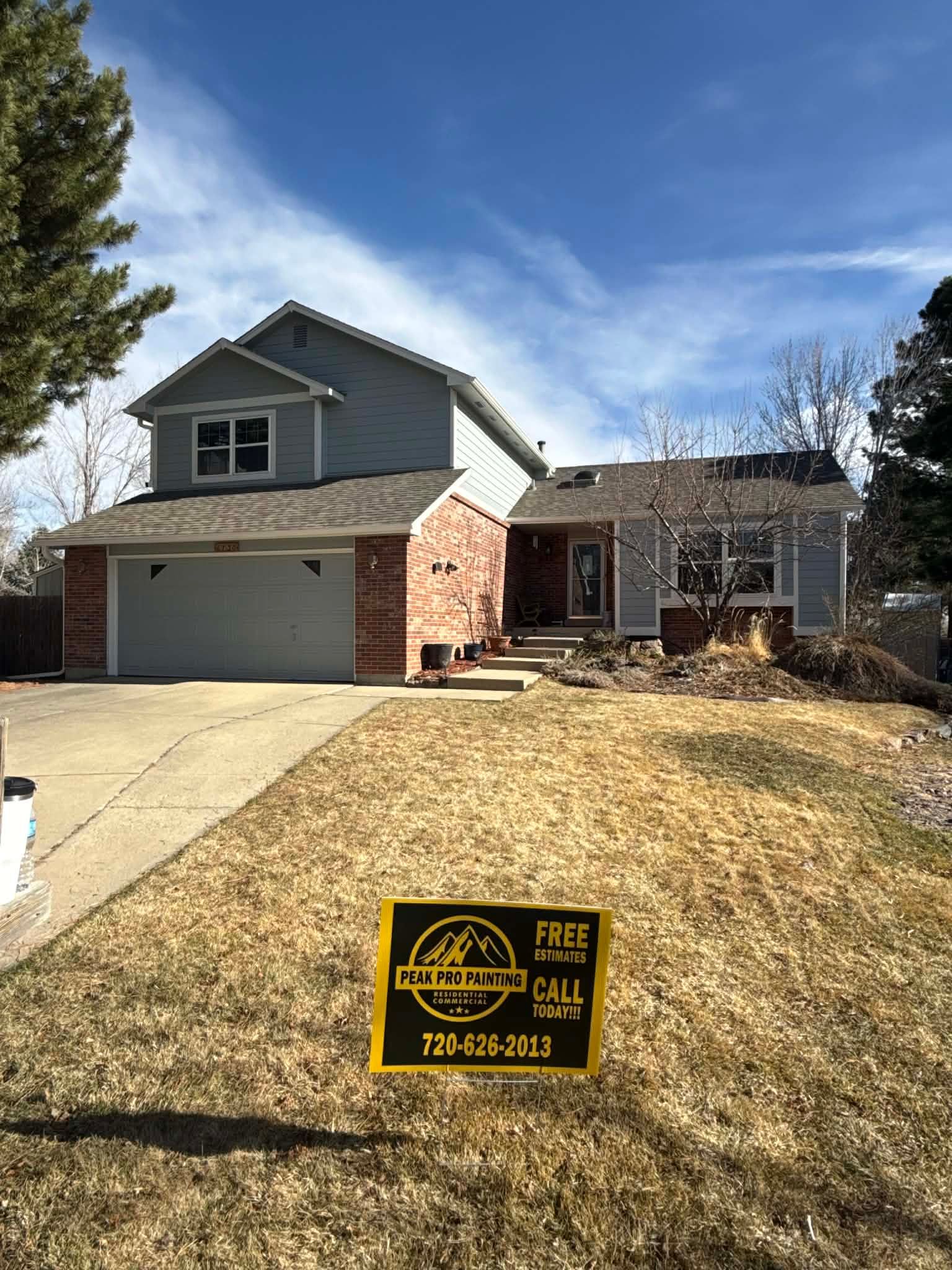 A two-story gray and brick suburban home with a garage, front yard, and a real estate sign in the grass.