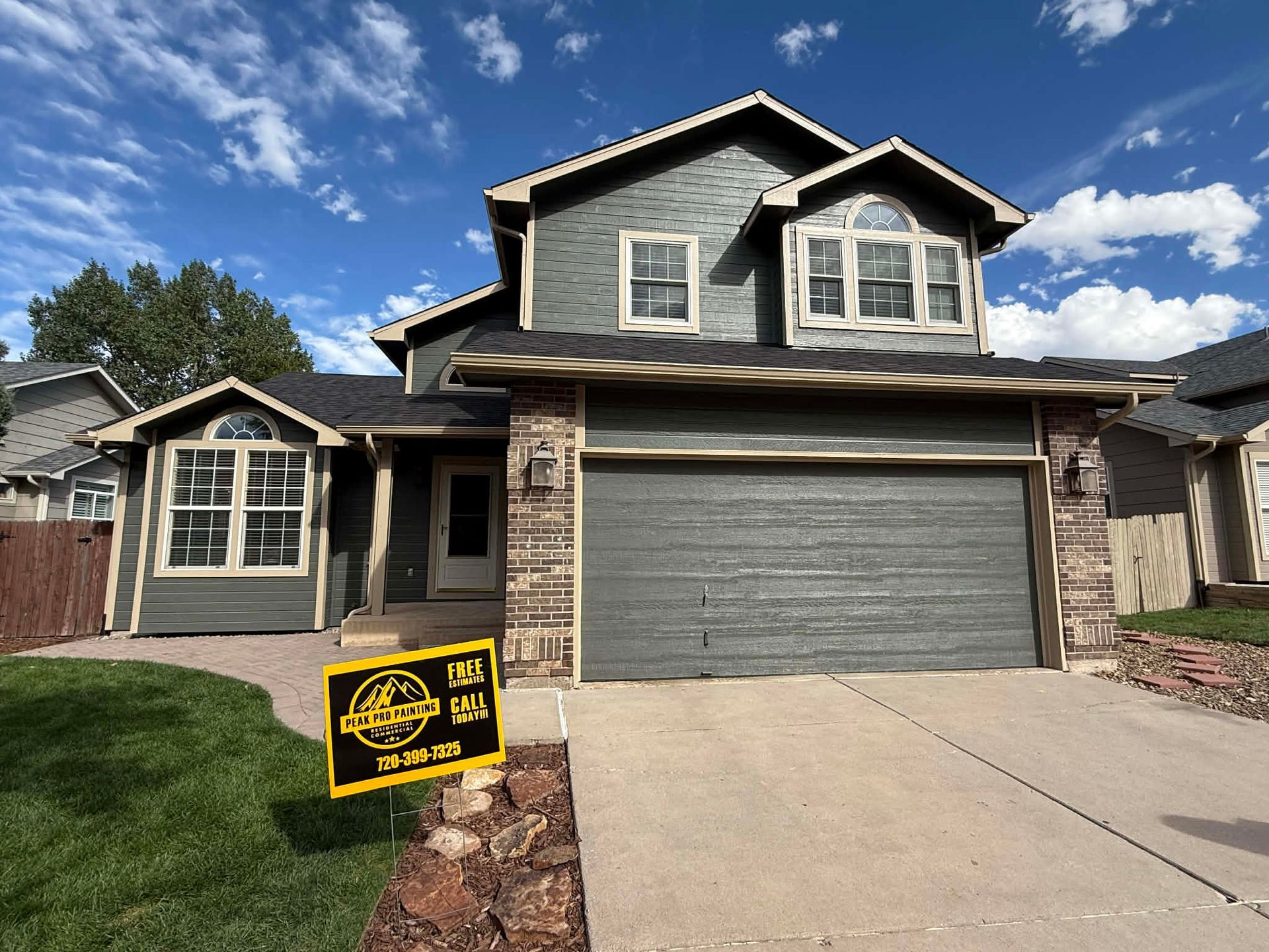 A two-story grey house with a stone facade, attached garage, and a yellow real estate sign on the front lawn.