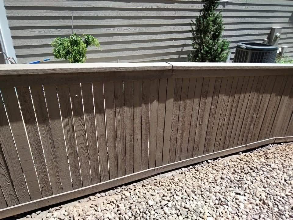 A light brown, vertical-slat privacy fence sits on a gravel ground next to a house siding with a small evergreen plant.
