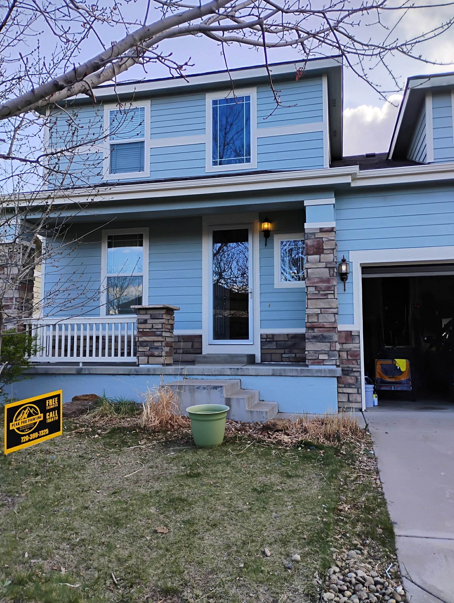 A light blue two-story house with stone accents, a front porch, and a partially open garage under a bare tree.