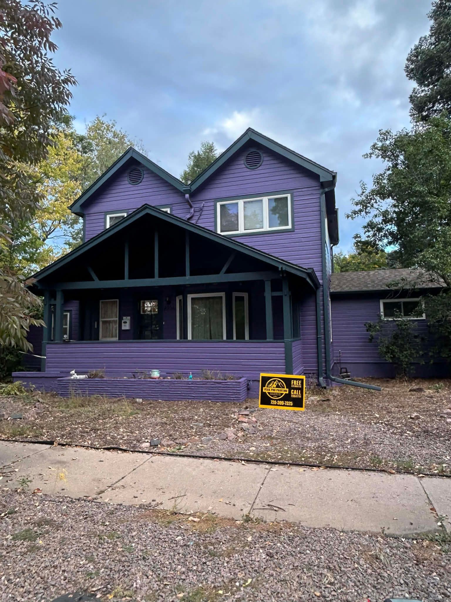 A two-story purple house with a large front porch and a dark roof, surrounded by trees on a cloudy day.