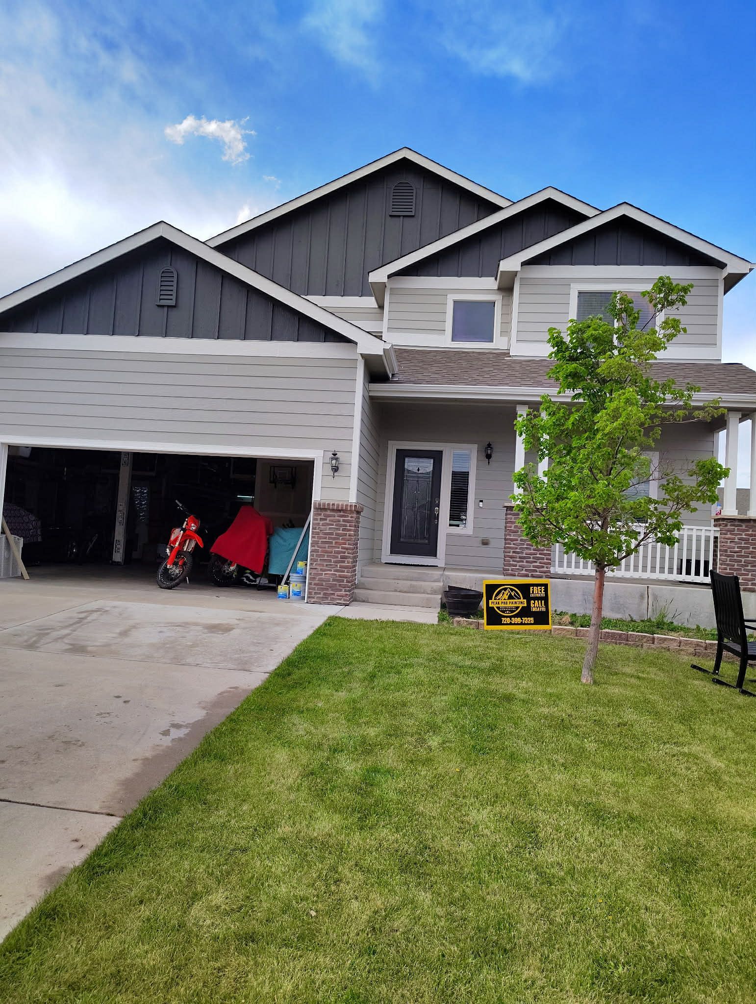 A two-story gray house with dark gables, a front porch, and an open garage containing a red vehicle under a blue sky.