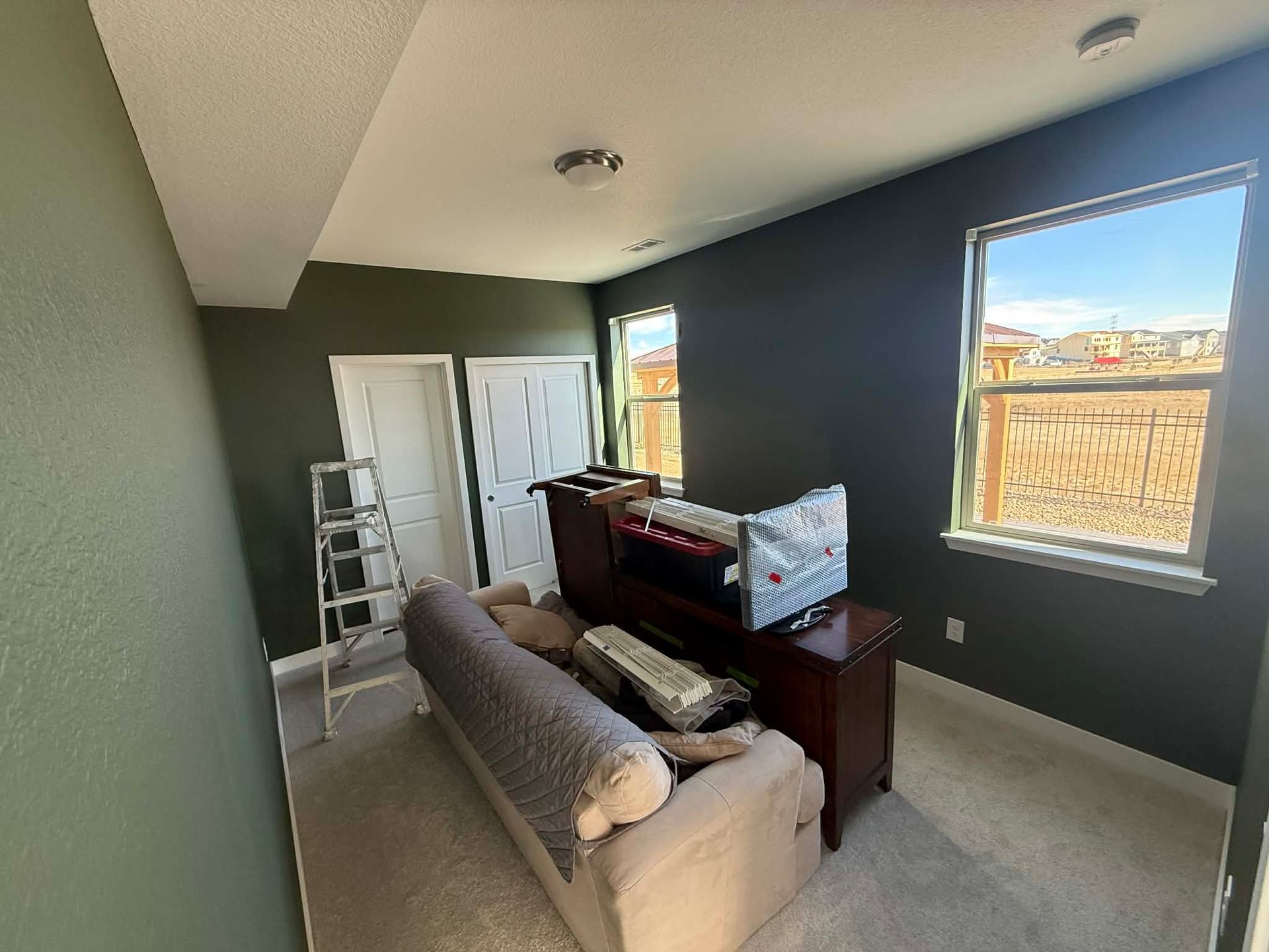 A room with dark green walls, a beige sofa facing a television on a wooden stand, white doors, a ladder, and a window.