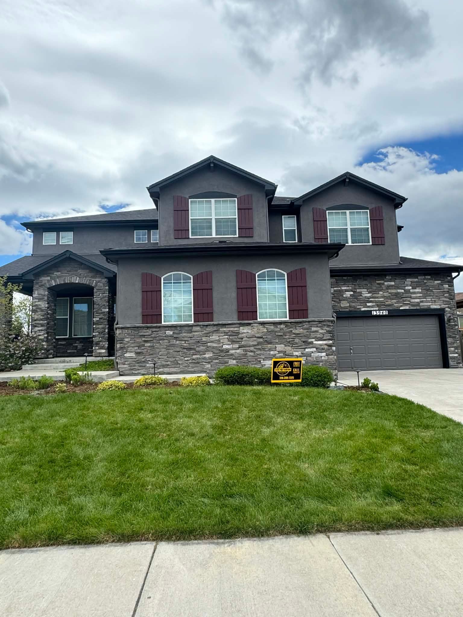 A two-story suburban house with grey stucco, brown shutters, stone facade, and an attached two-car garage.