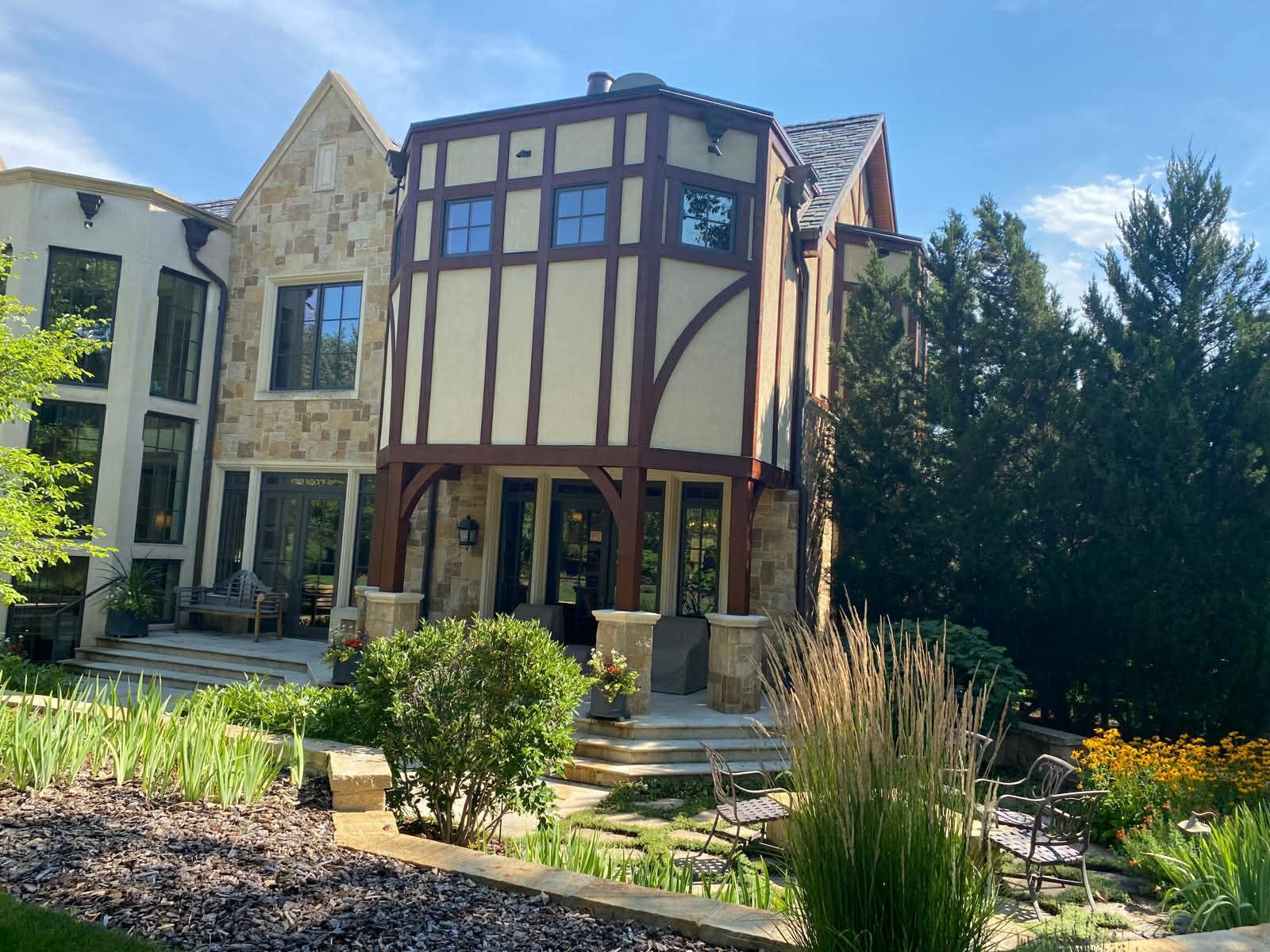 A two-story home with tan stucco, dark timber framing, and stone accents, surrounded by lush landscaping under a blue sky.