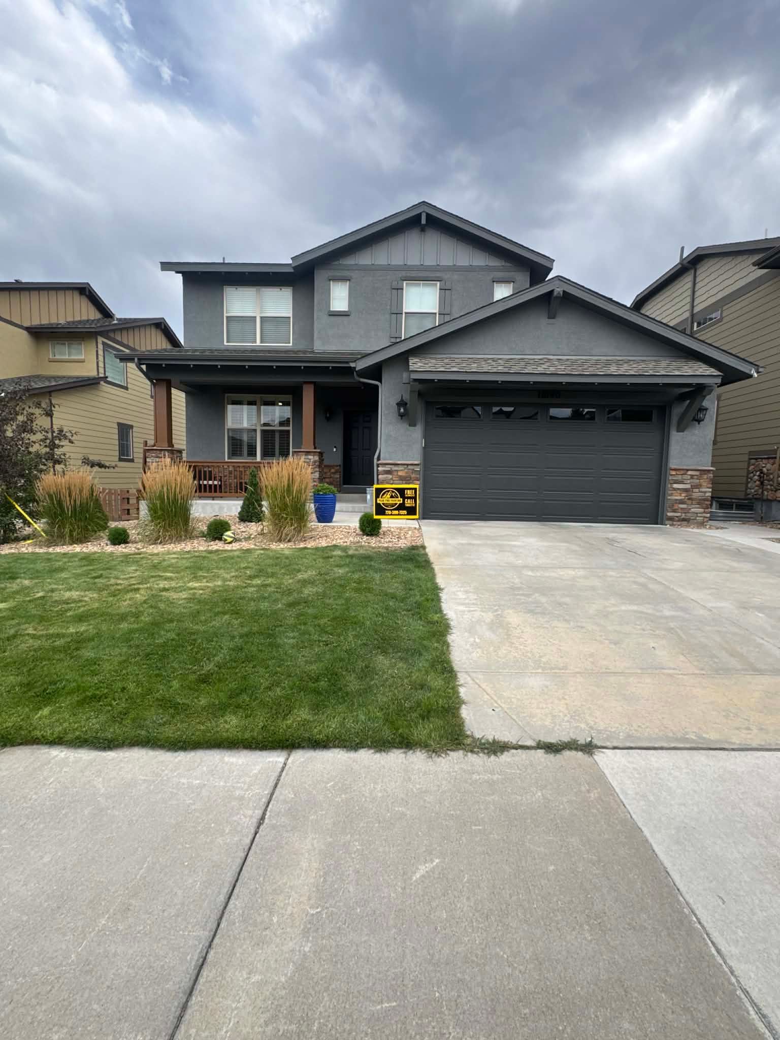 A two-story grey suburban house with a front lawn, porch, and attached garage under a cloudy sky.