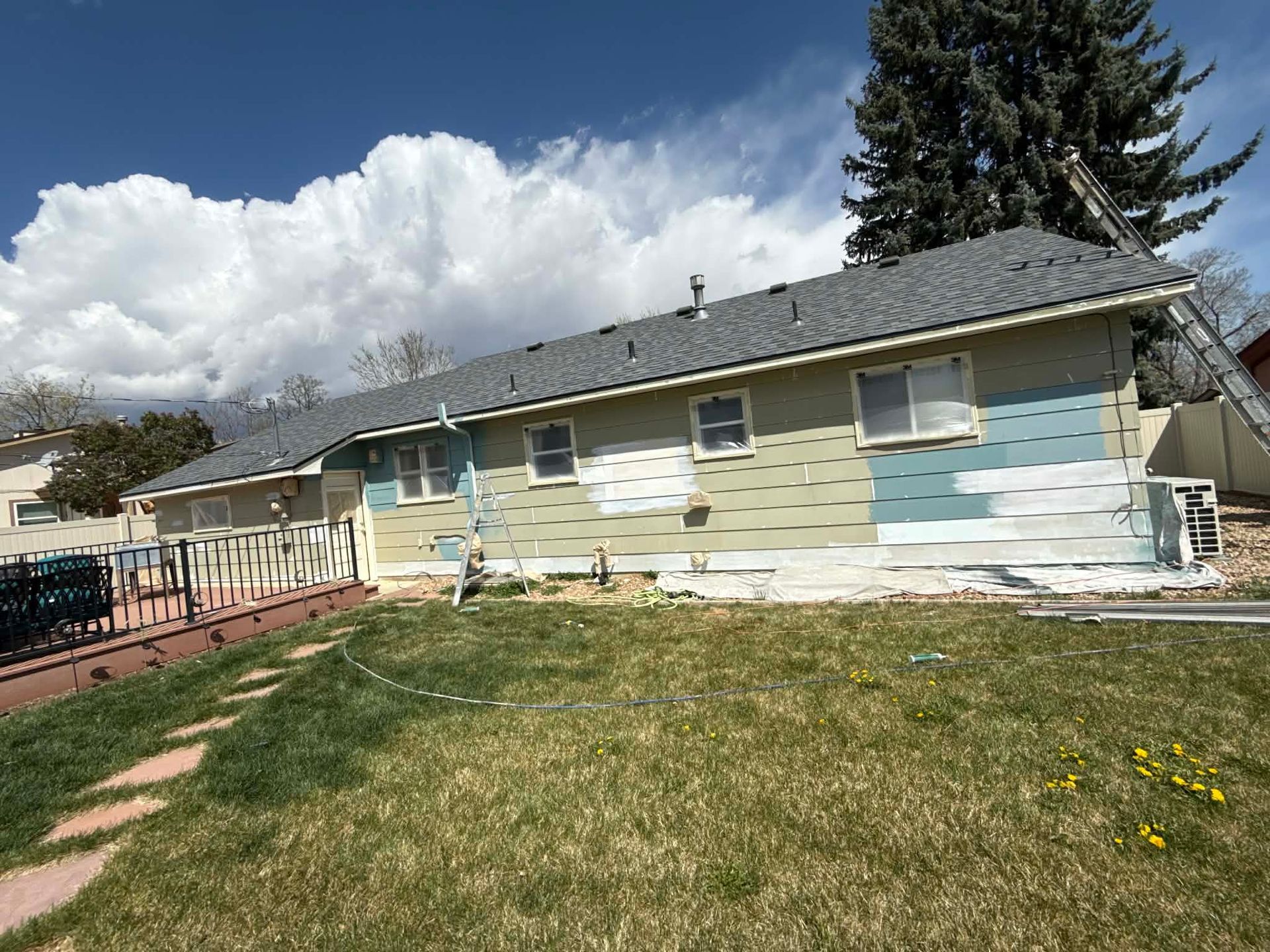 A house exterior undergoing renovation with patches of new siding and paint, featuring a roof, lawn, and a ladder.
