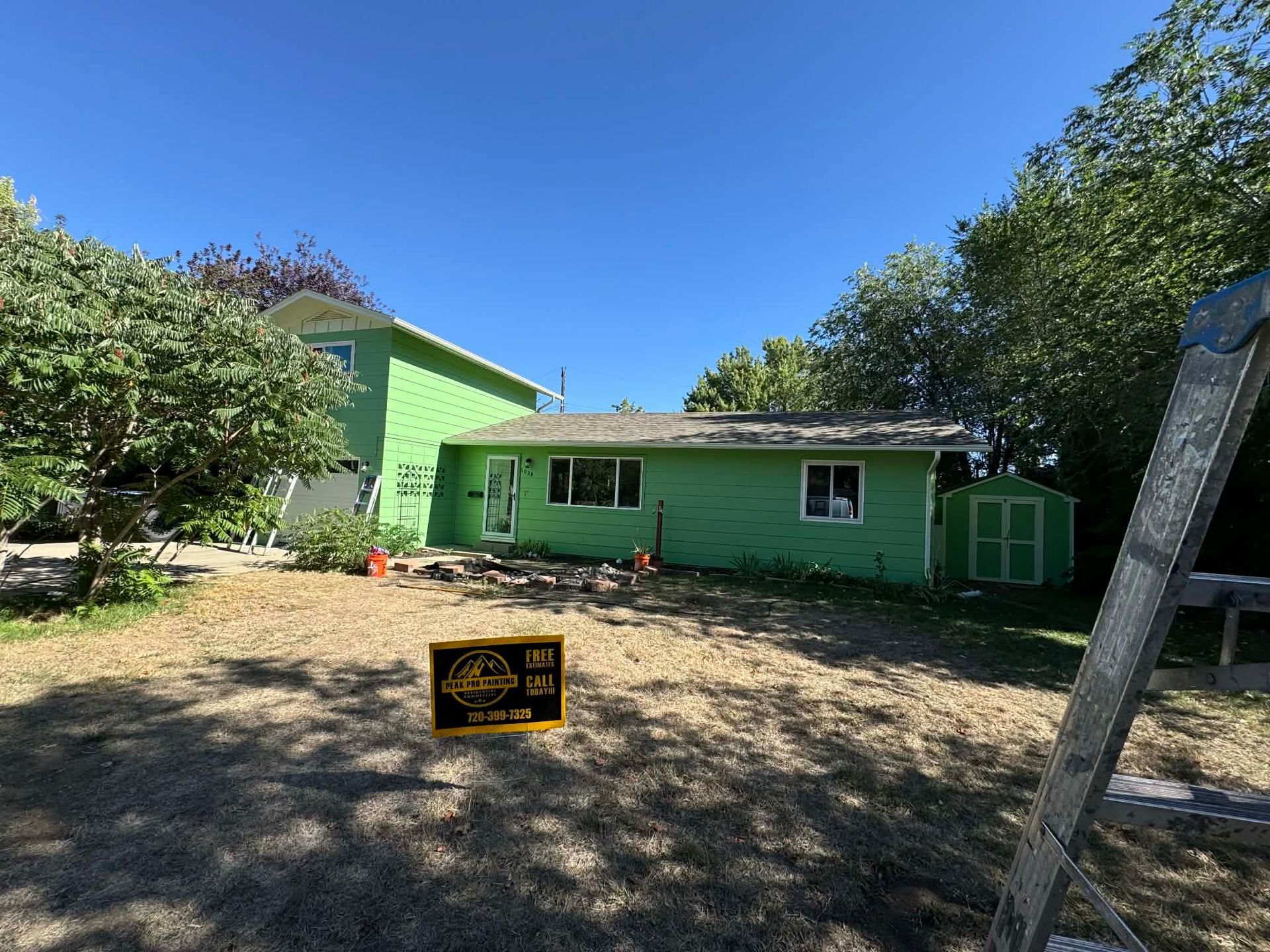 A bright green, two-story house with a small shed under a clear blue sky, seen from a lawn with a yellow sign.