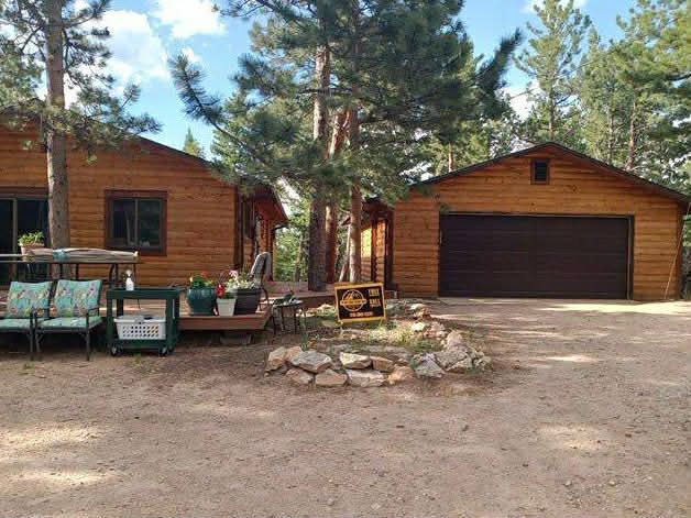 A wooden cabin with a matching garage, patio furniture, and a gravel driveway in a pine tree forest.
