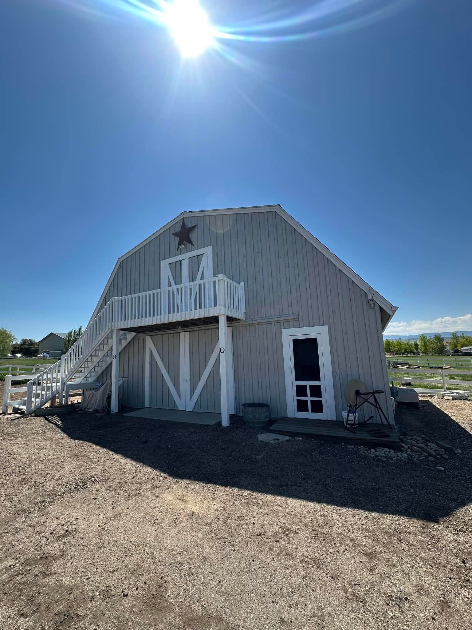 A gray barn with a white balcony and door under a bright sun, set on a gravel lot under a clear blue sky.