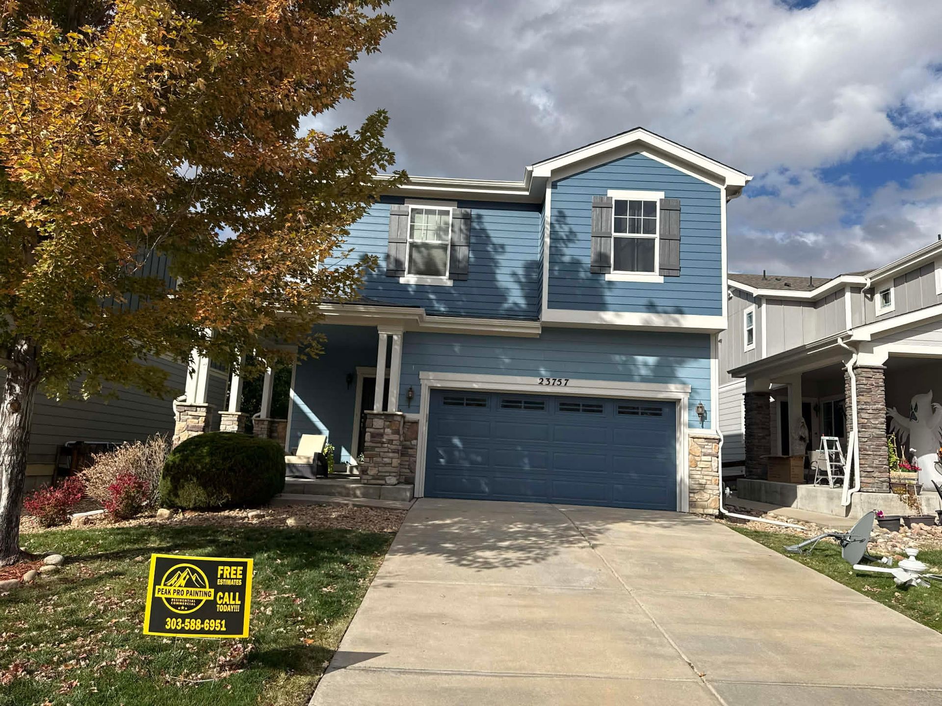 A two-story blue house with a gray garage, stone accents, and a yellow sign on the front lawn under a cloudy sky.