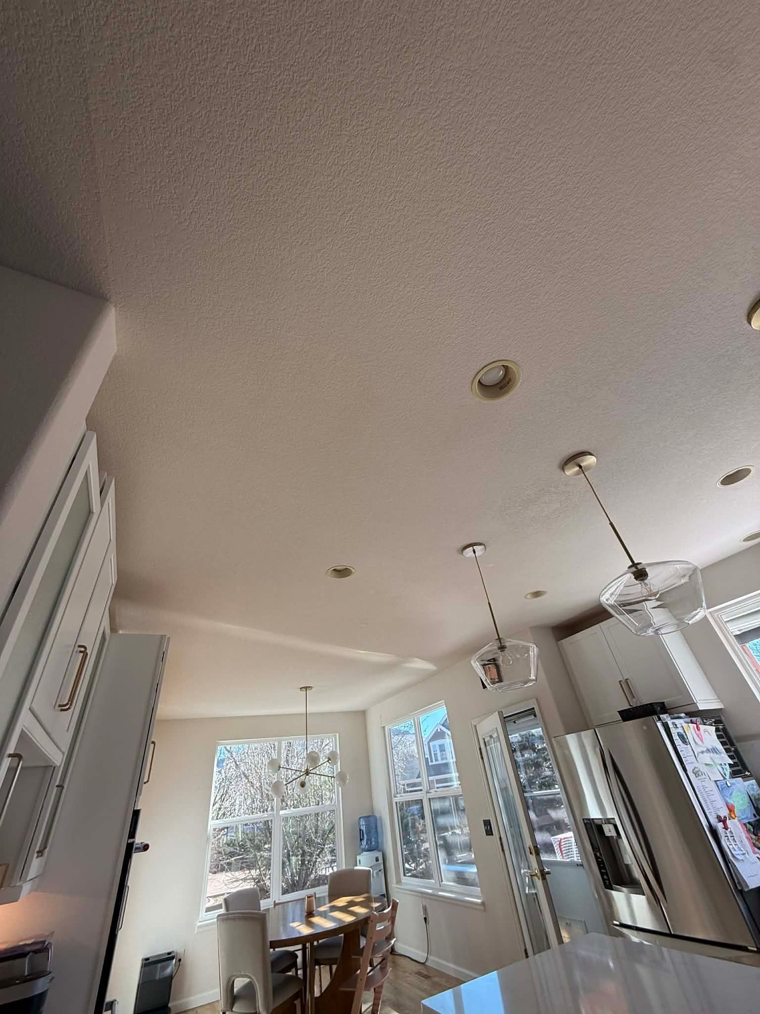 A modern, sunlit kitchen features white cabinets, pendant lighting, recessed lights, and a dining area by large windows.