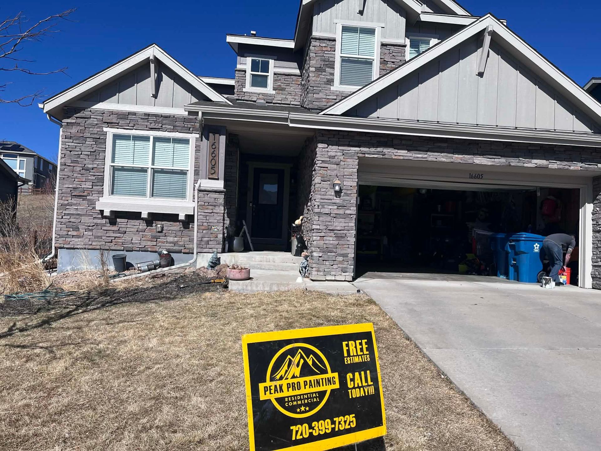 A two-story stone-front house with a garage and a yard sign in the foreground under a clear blue sky.