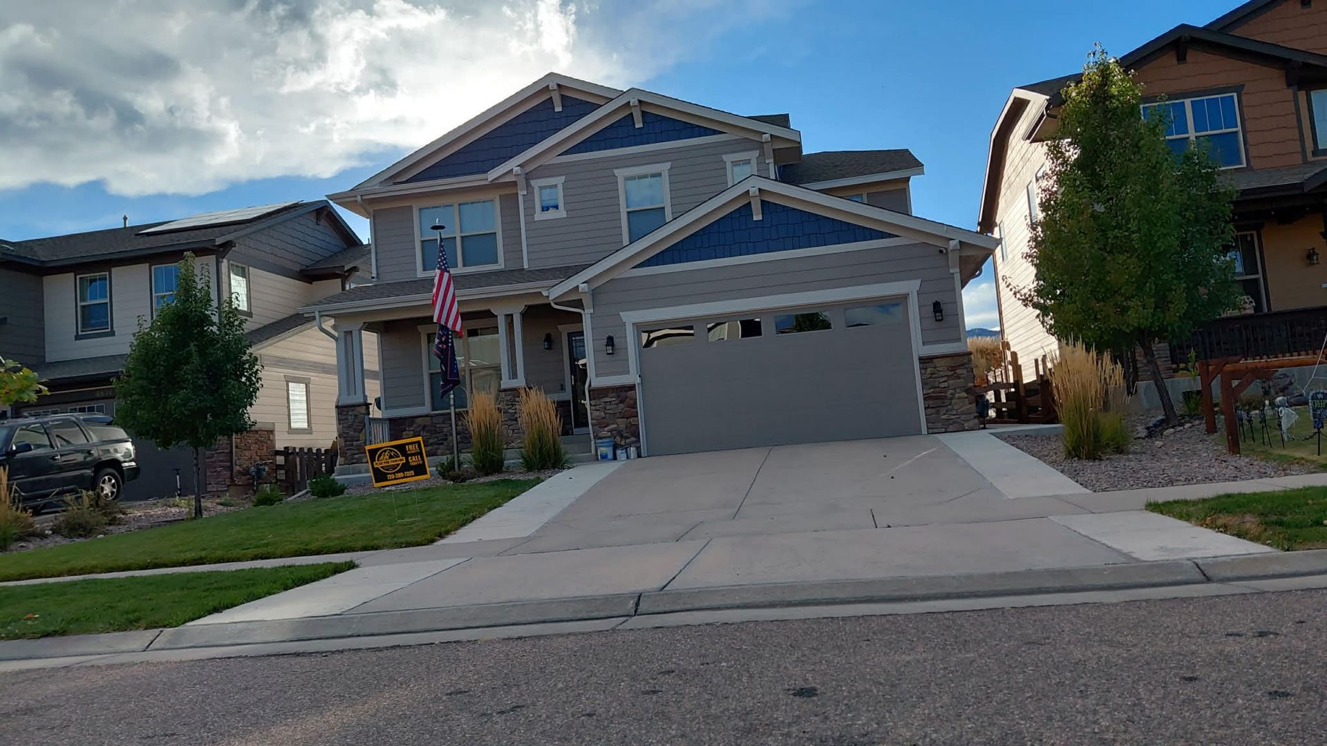 A multi-story gray residential house with a blue-accented roof, front porch, two-car garage, and American flag.