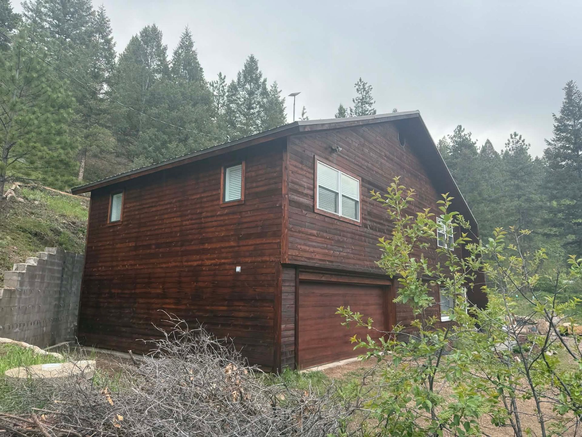A dark-brown, two-story wooden cabin with a garage sits nestled against a hillside covered in pine trees.