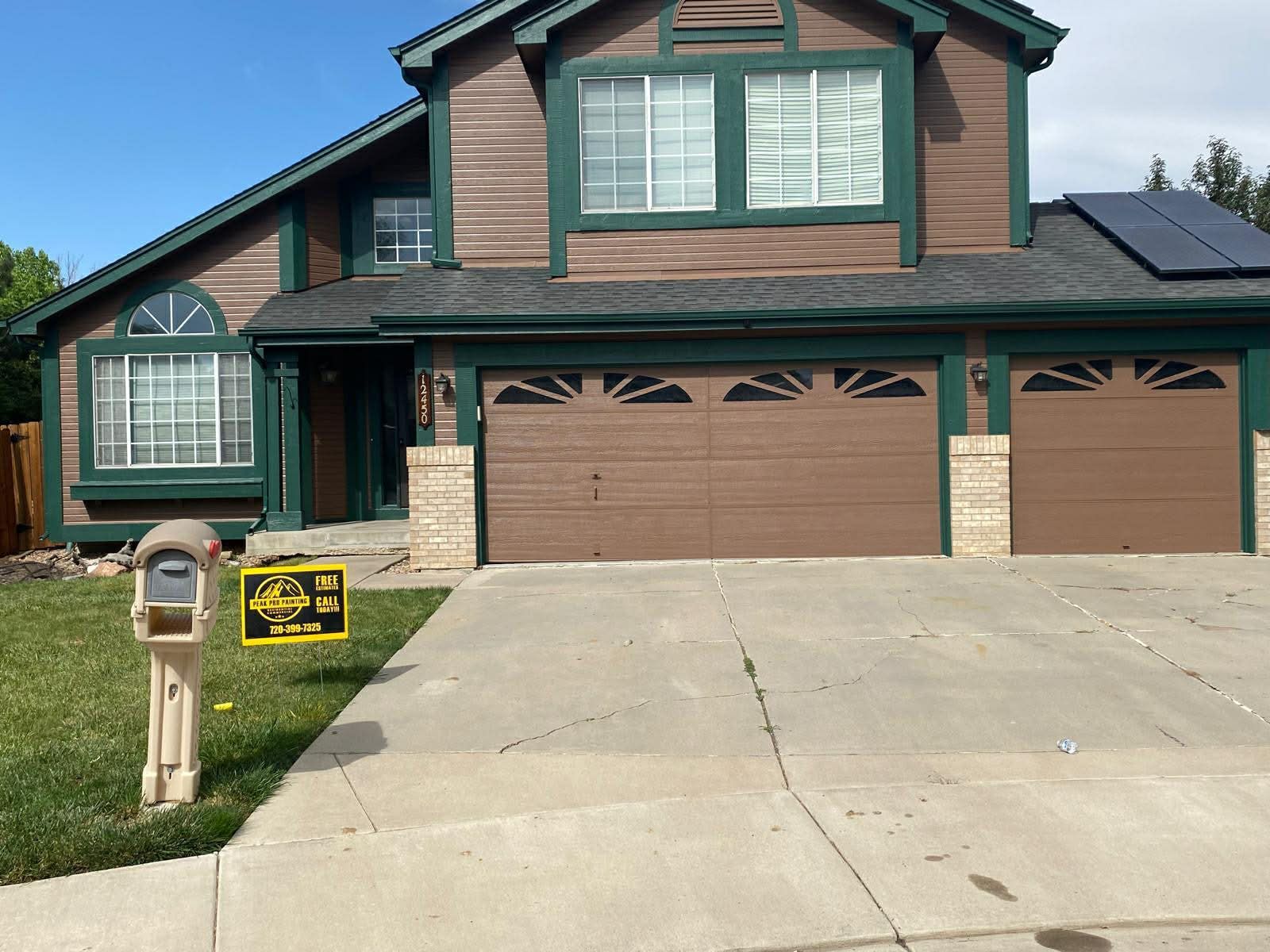 A two-story brown house with green trim, a three-car garage, a concrete driveway, and a mailbox on the front lawn.