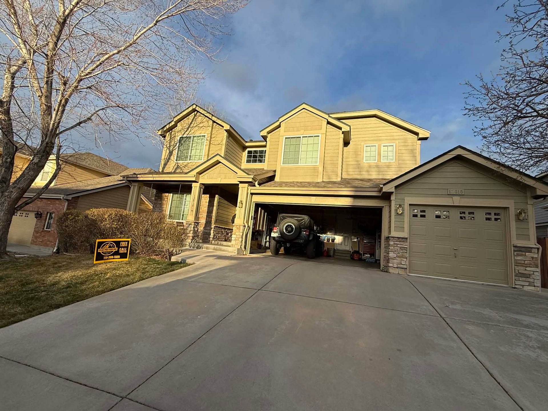 A two-story tan suburban house with a paved driveway, a garage, and an SUV parked under an open carport area.