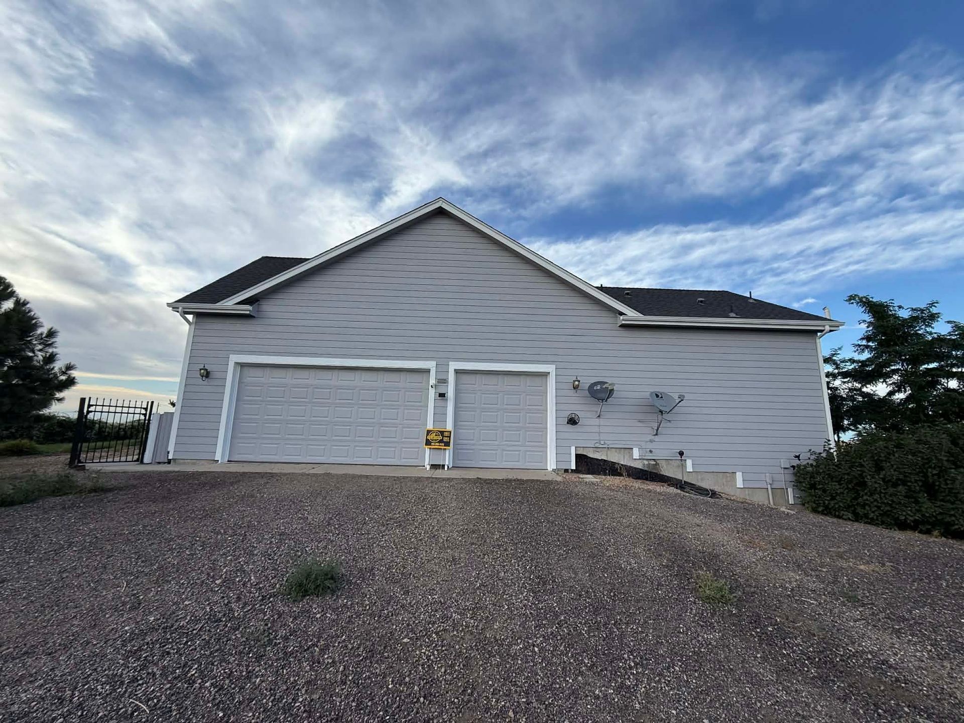 A detached light-gray garage with a dark shingled roof under a blue, cloudy sky, set on a gravel lot.